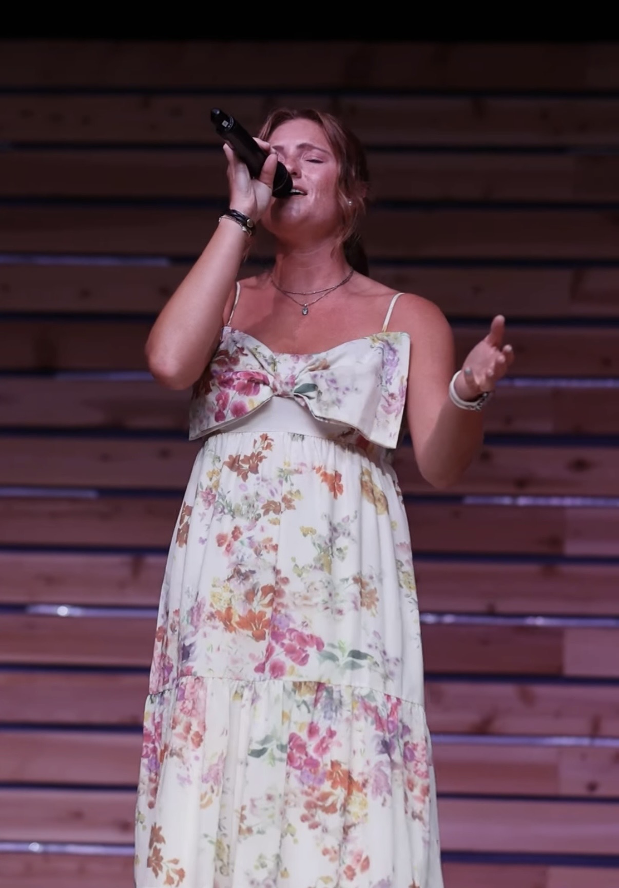 Woman singing into a microphone on stage with wooden background, wearing a floral dress with spaghetti straps.