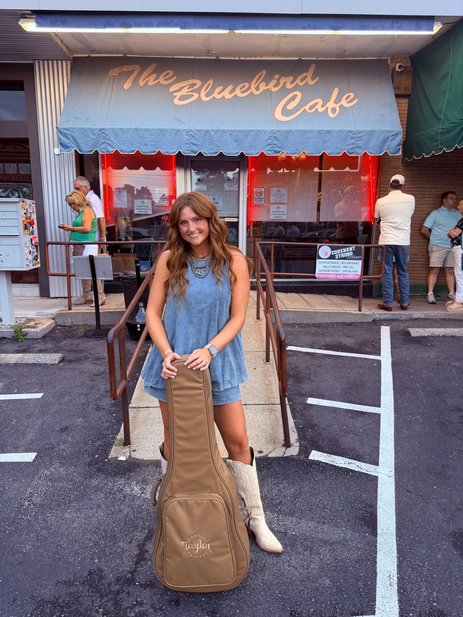 Young woman in a blue dress holding a guitar case, standing in front of a cafe entrance with a blue awning.