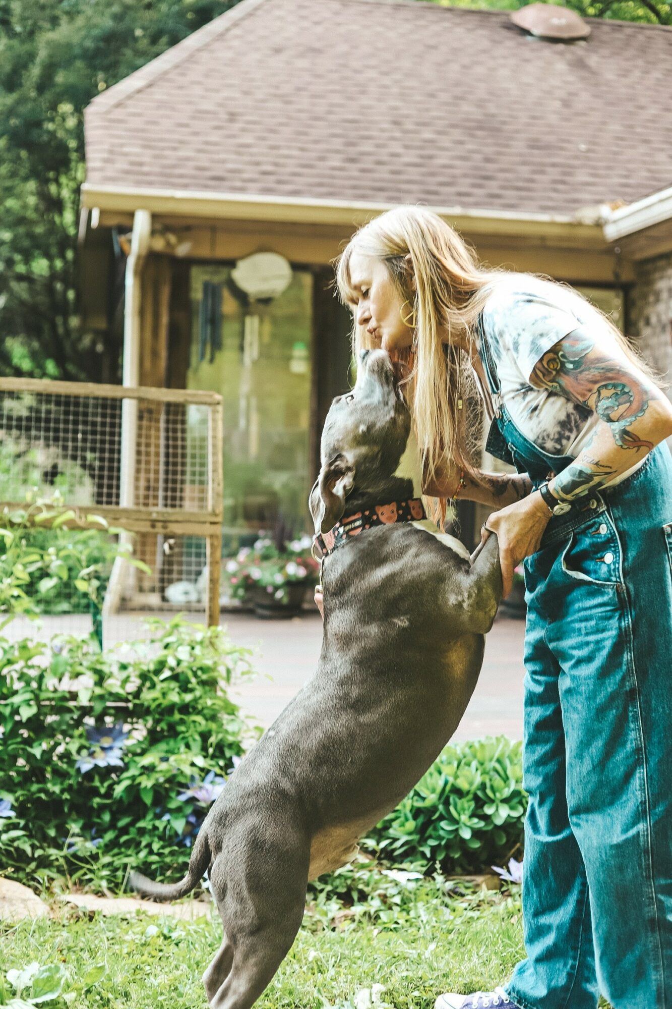 Woman with long hair and tattoos interacts with a jumping dog outdoors in a garden area.