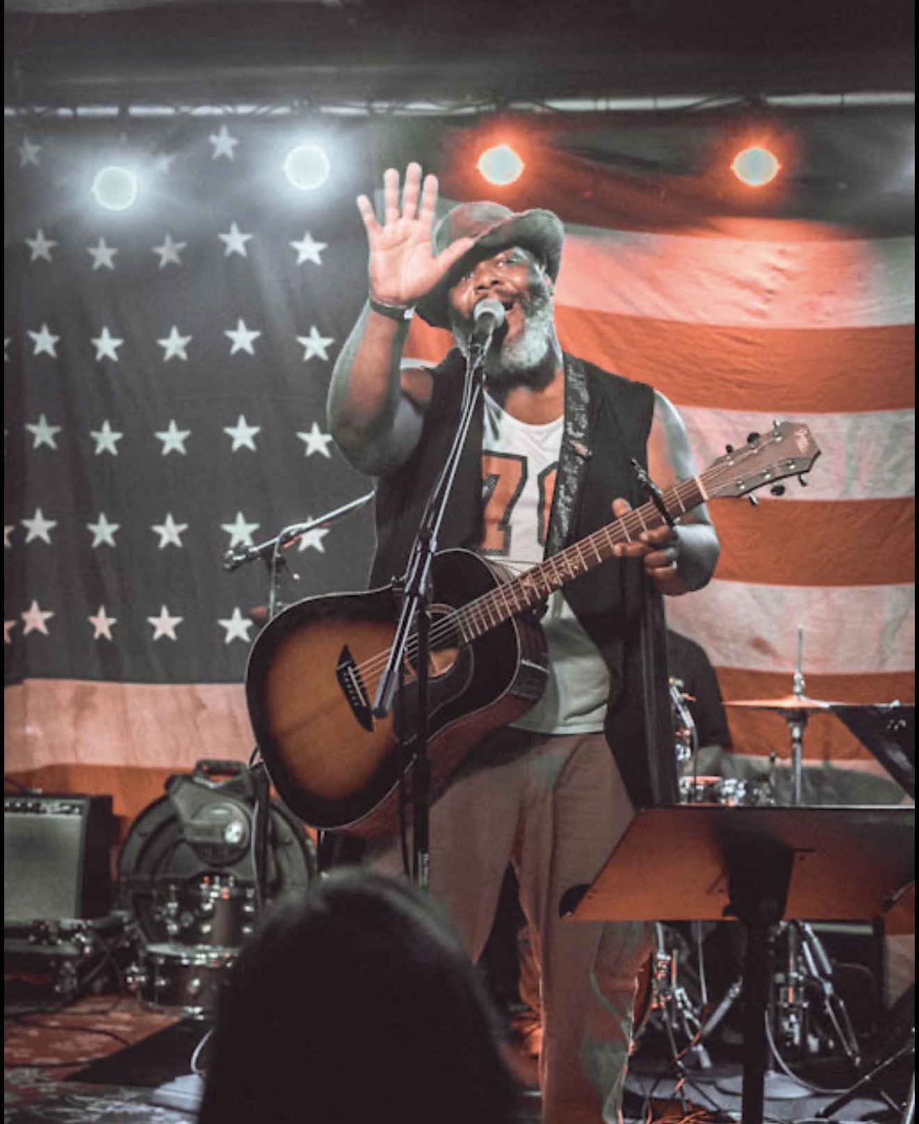 Man with a beard and glasses singing and playing guitar on stage with American flag backdrop, raising his hand.
