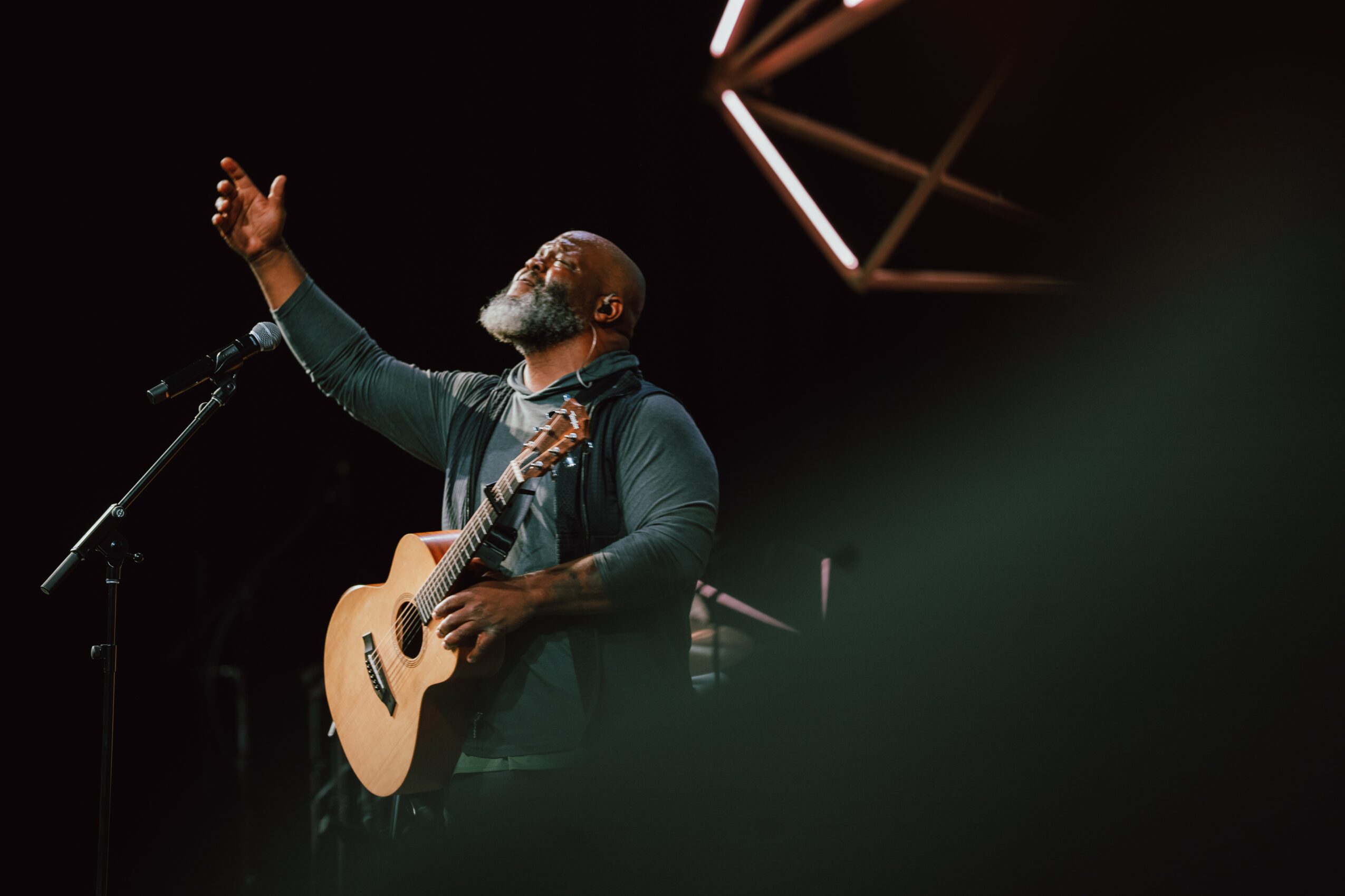 Man with gray beard and glasses singing with a guitar on stage, raising one hand, with stage lighting in background.