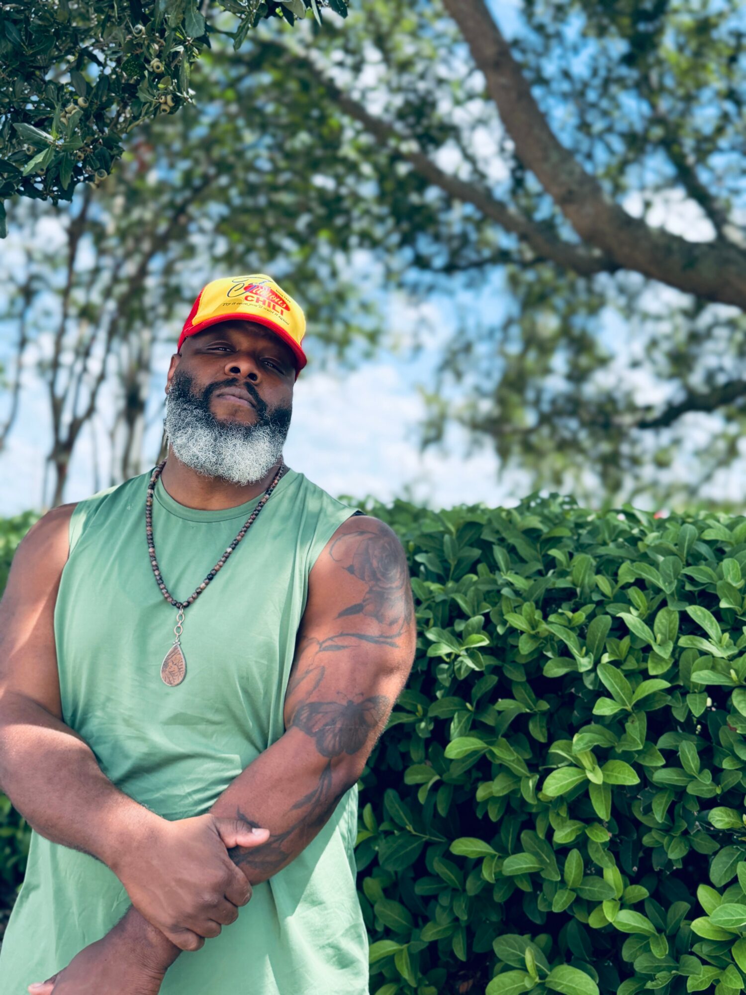 Man with a beard and tattoos wearing a green sleeveless shirt and a red cap outdoors, standing next to a bush.
