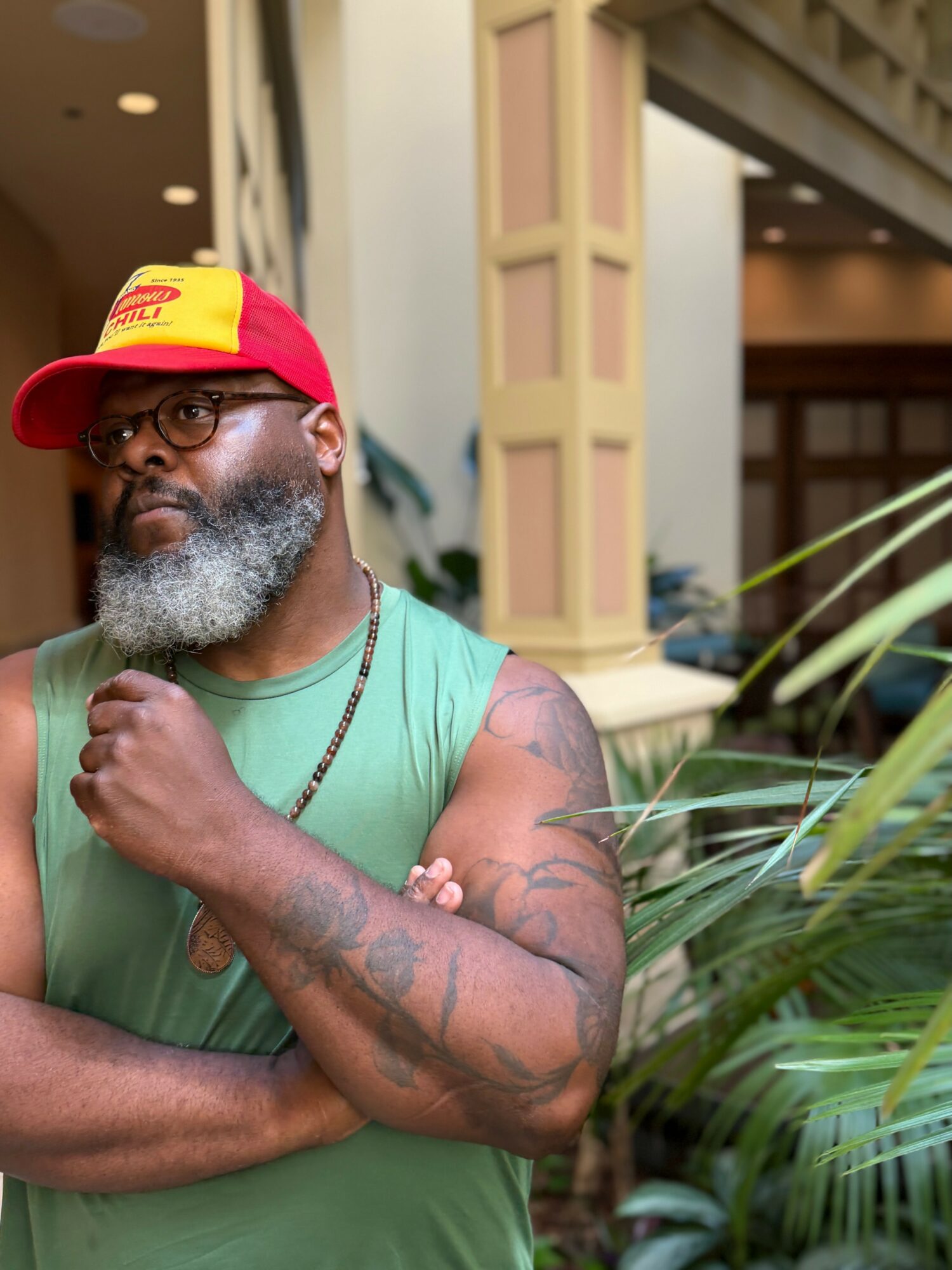 Man with glasses, beard, and tattoos wearing a red and yellow cap and green sleeveless shirt, standing outdoors.