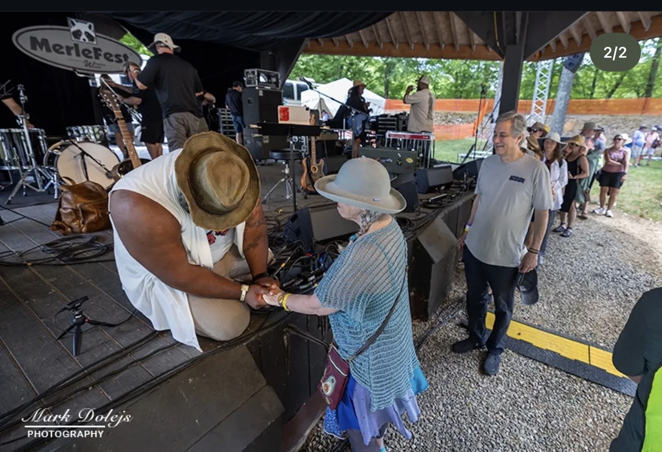 Two women exchanging items at an outdoor event under a tent, with a band playing in the background and people nearby.