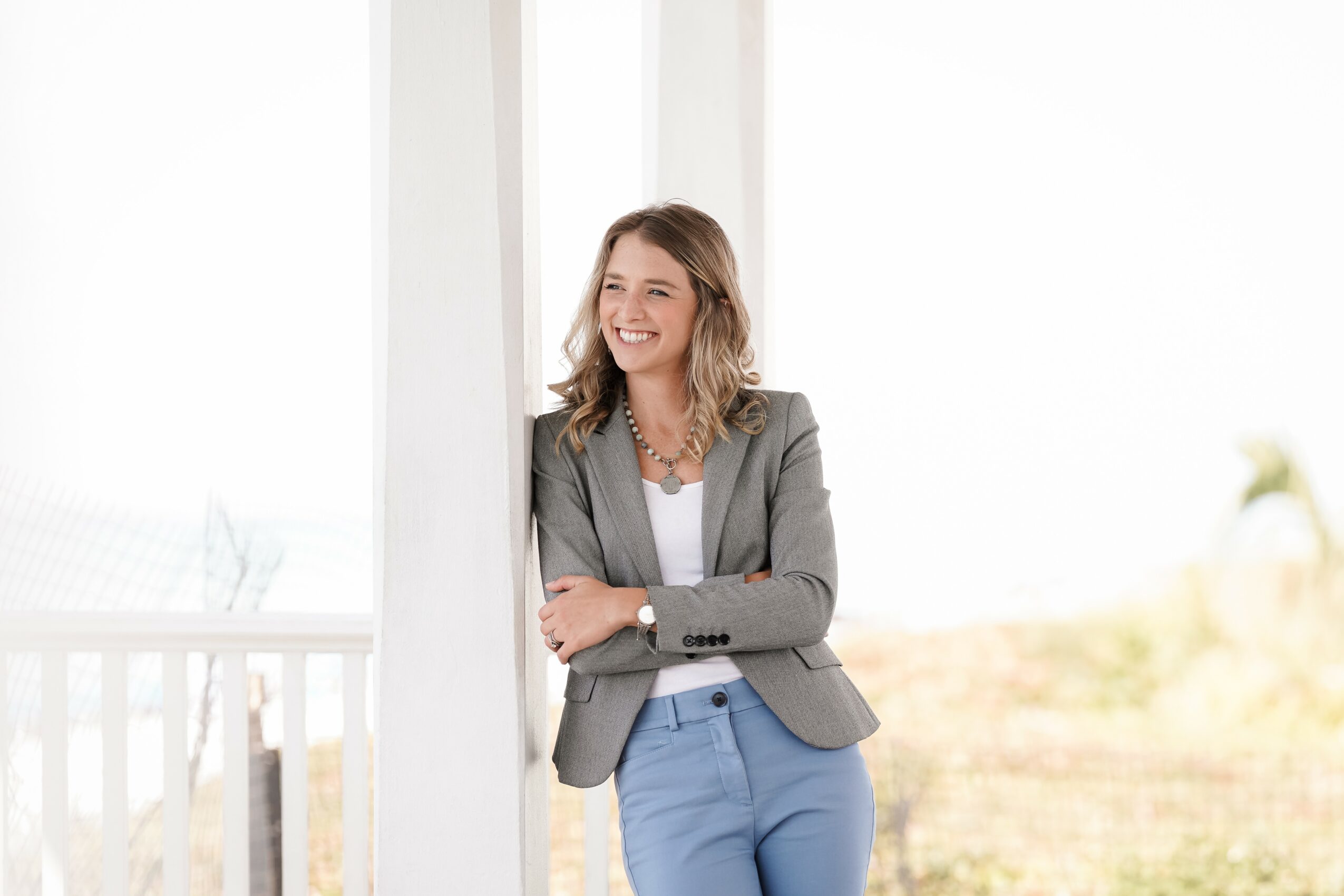 Woman with shoulder-length hair smiling, wearing a gray blazer and blue pants, standing near a window with sunlight.