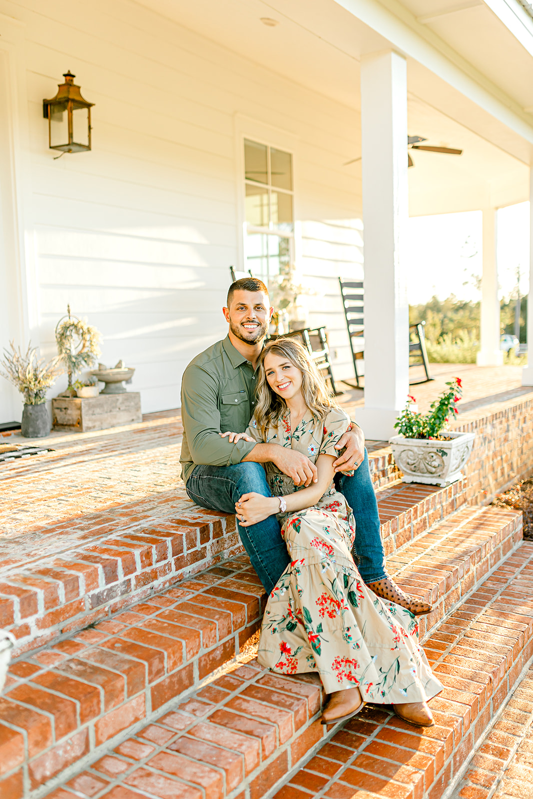 A smiling man and woman sit on brick steps outside a house, with sunlight and greenery in the background.