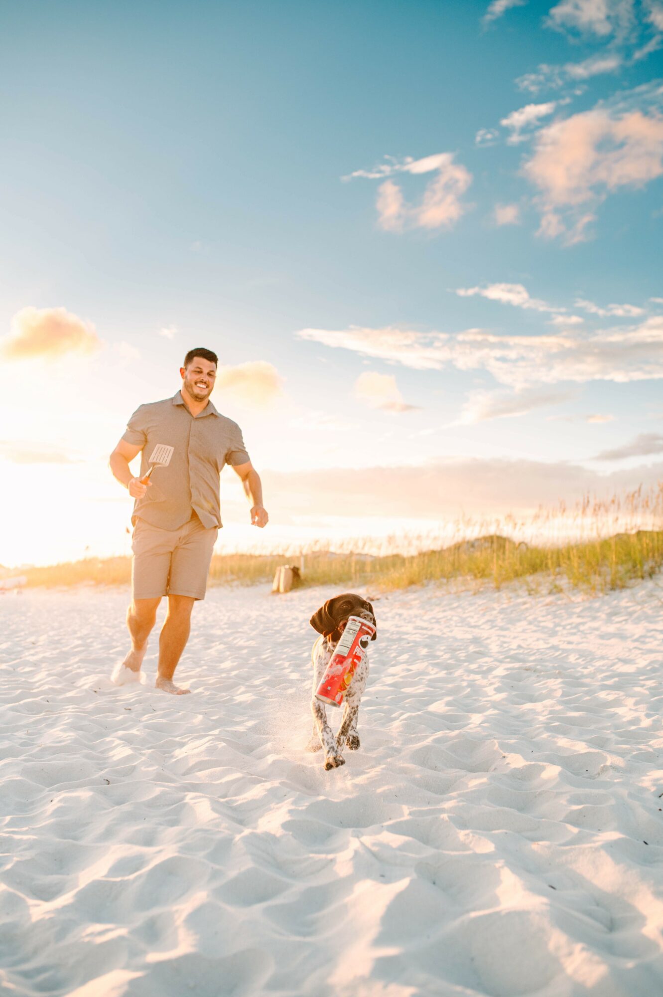 Man and child playing on sandy beach with blue sky and clouds, sunlight shining, grassy dunes in background.