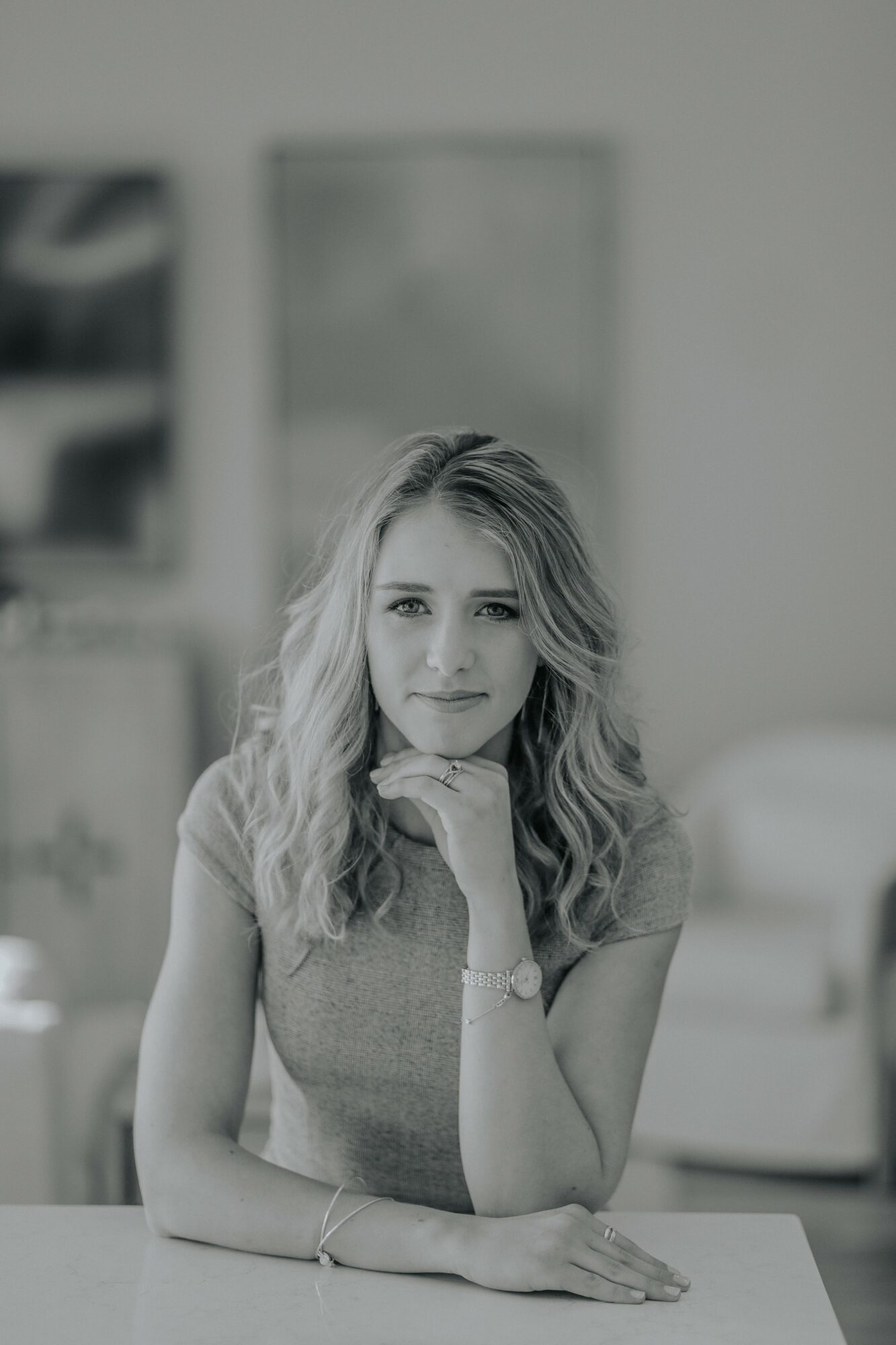 Young woman with wavy hair resting her chin on her hand, sitting at a table in a room.