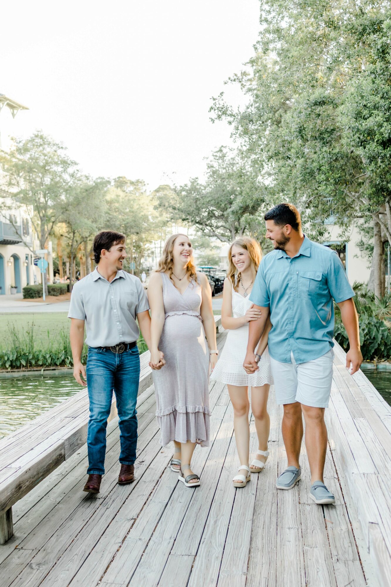 Four people walk on a wooden path near water, trees, and buildings in the background, holding hands and talking.