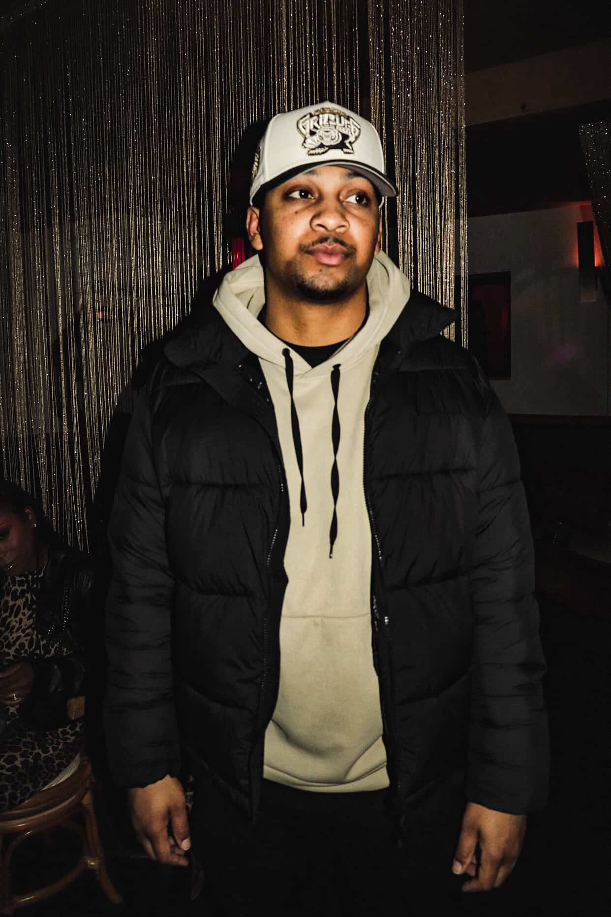 Young man wearing a baseball cap, hoodie, and jacket standing indoors with dark background.