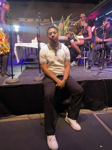 Young man sitting on stage with musical performers in background, stage setup, and plants, facing forward.