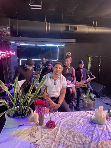 Group of musicians performing on stage with a person sitting at a table in front, decorated with flowers and candles.