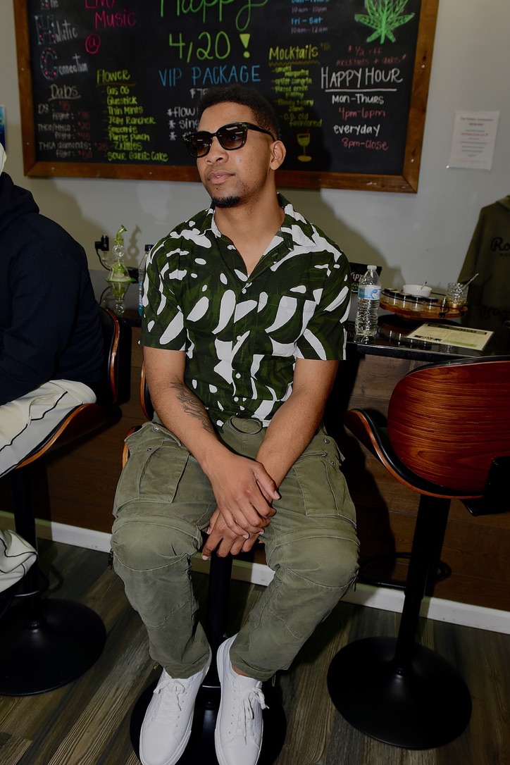 Man wearing sunglasses, patterned shirt, and khaki pants sitting at a bar or cafe, with a blackboard menu behind him.