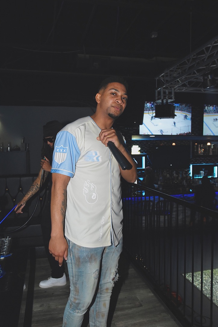 Young man in a light-colored sports jersey and jeans standing indoors with a dark background and stage lighting.