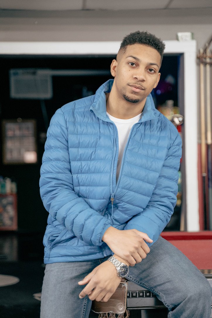 Young man with short curly hair wearing a blue puffer jacket and gray pants, sitting with arms crossed in a room.