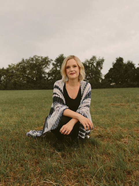 Woman sitting on grass in a park with trees in the background, smiling, wearing a patterned shawl and black clothing.