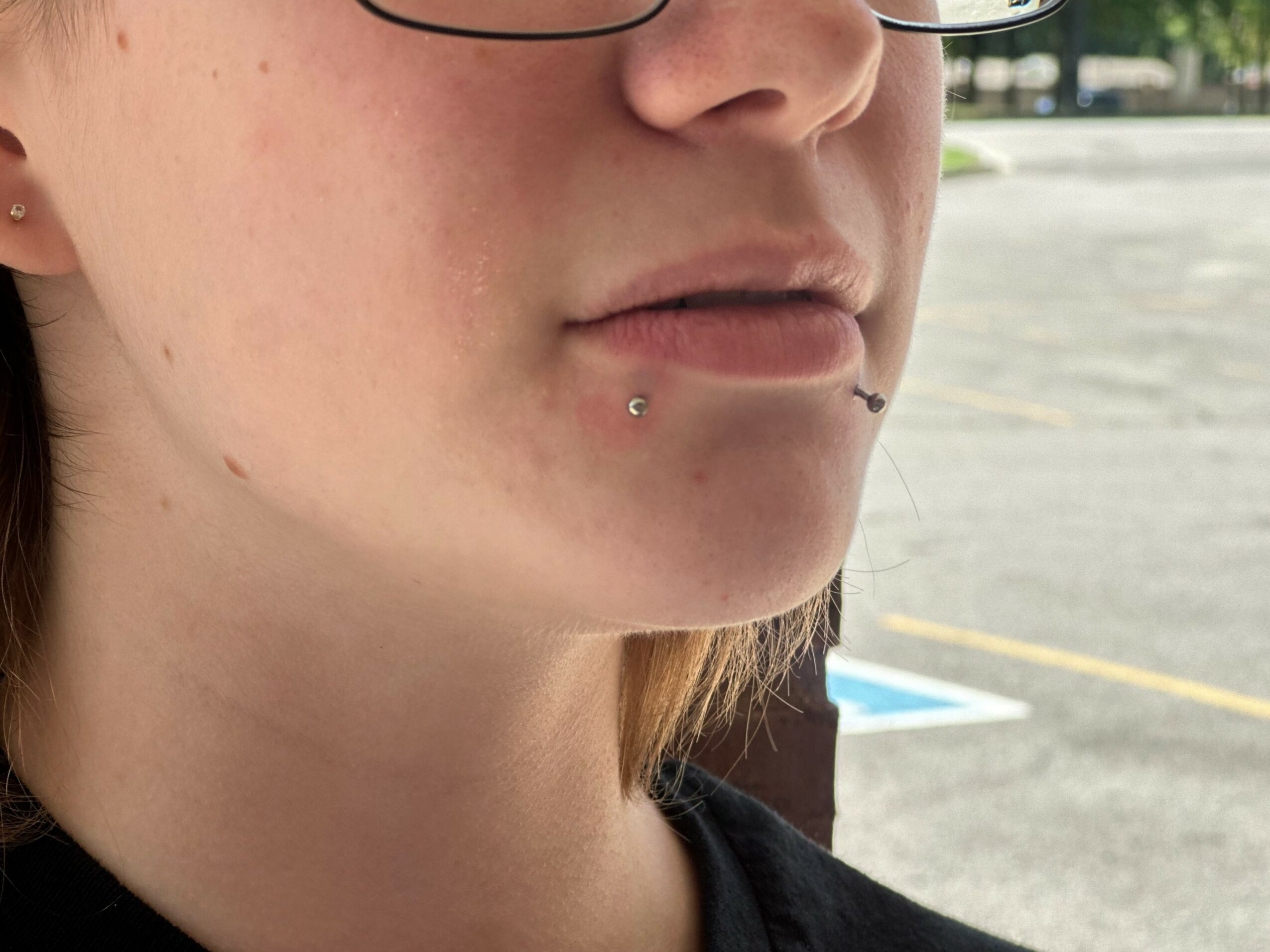 Close-up of a person's face showing lips, chin, and piercings, with a parking lot background.