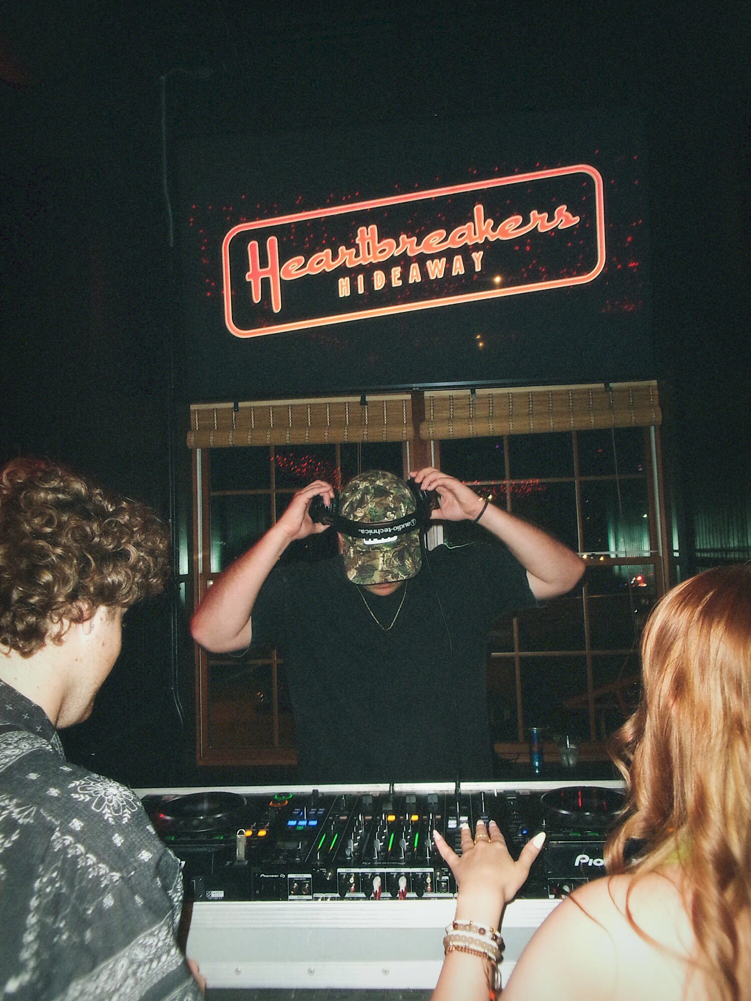 Person wearing a cap and headphones behind a DJ booth, with two people watching, and a sign reading 'Headquarters Highway' in the background.