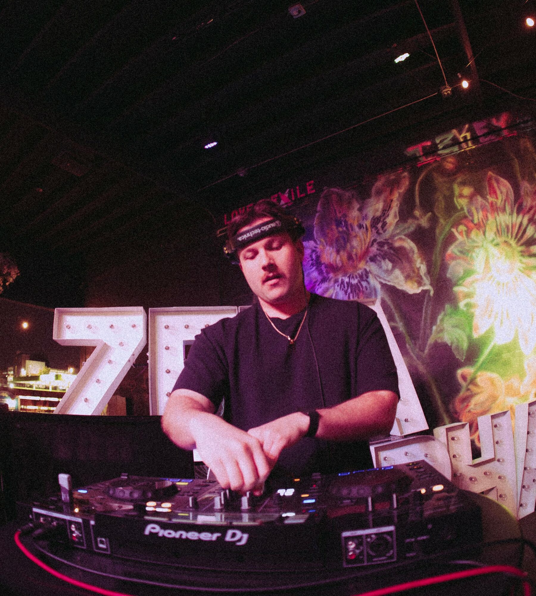 DJ with headphones operating turntables in a colorful, floral-lit setting, with illuminated letters in the background.