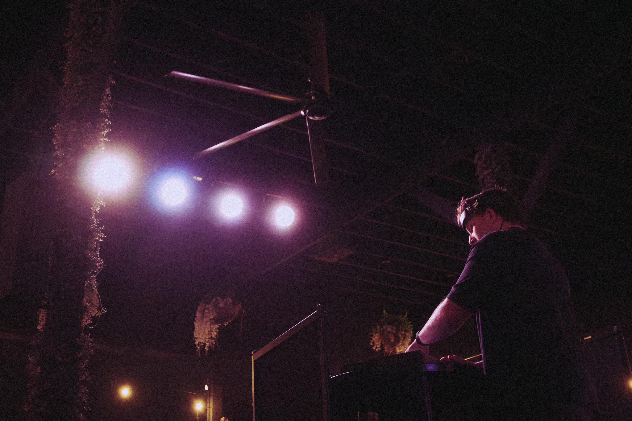 Person standing at a DJ booth with purple and white stage lights overhead, dark background, and ceiling visible.