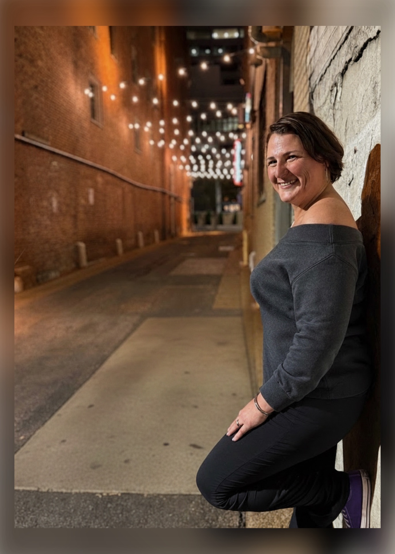 Smiling woman with short hair leaning against a wall in an urban alley at night, illuminated by string lights.