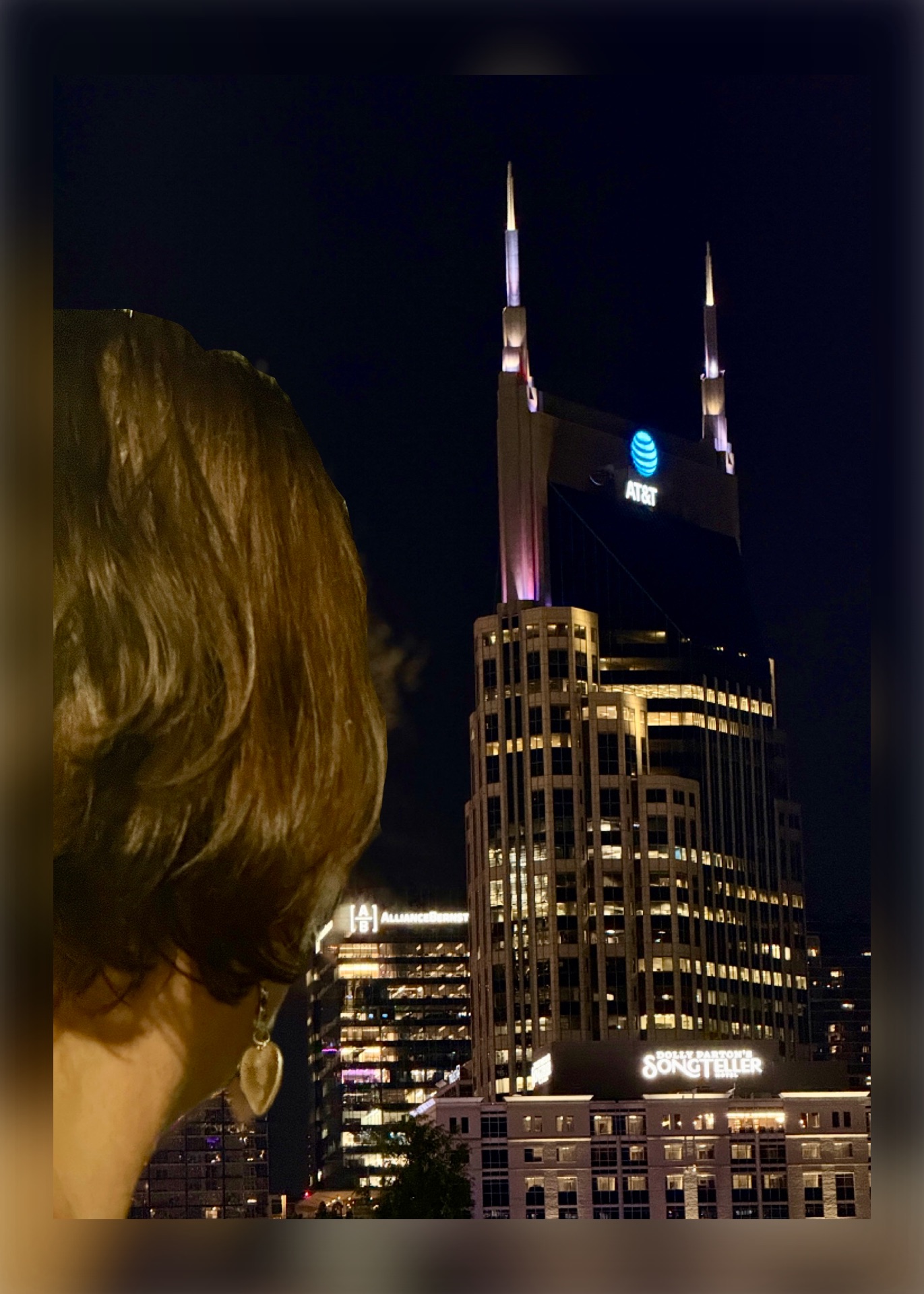 Nighttime cityscape with a tall building featuring illuminated spires and a blue logo, partially obscured by a person's head in the foreground.