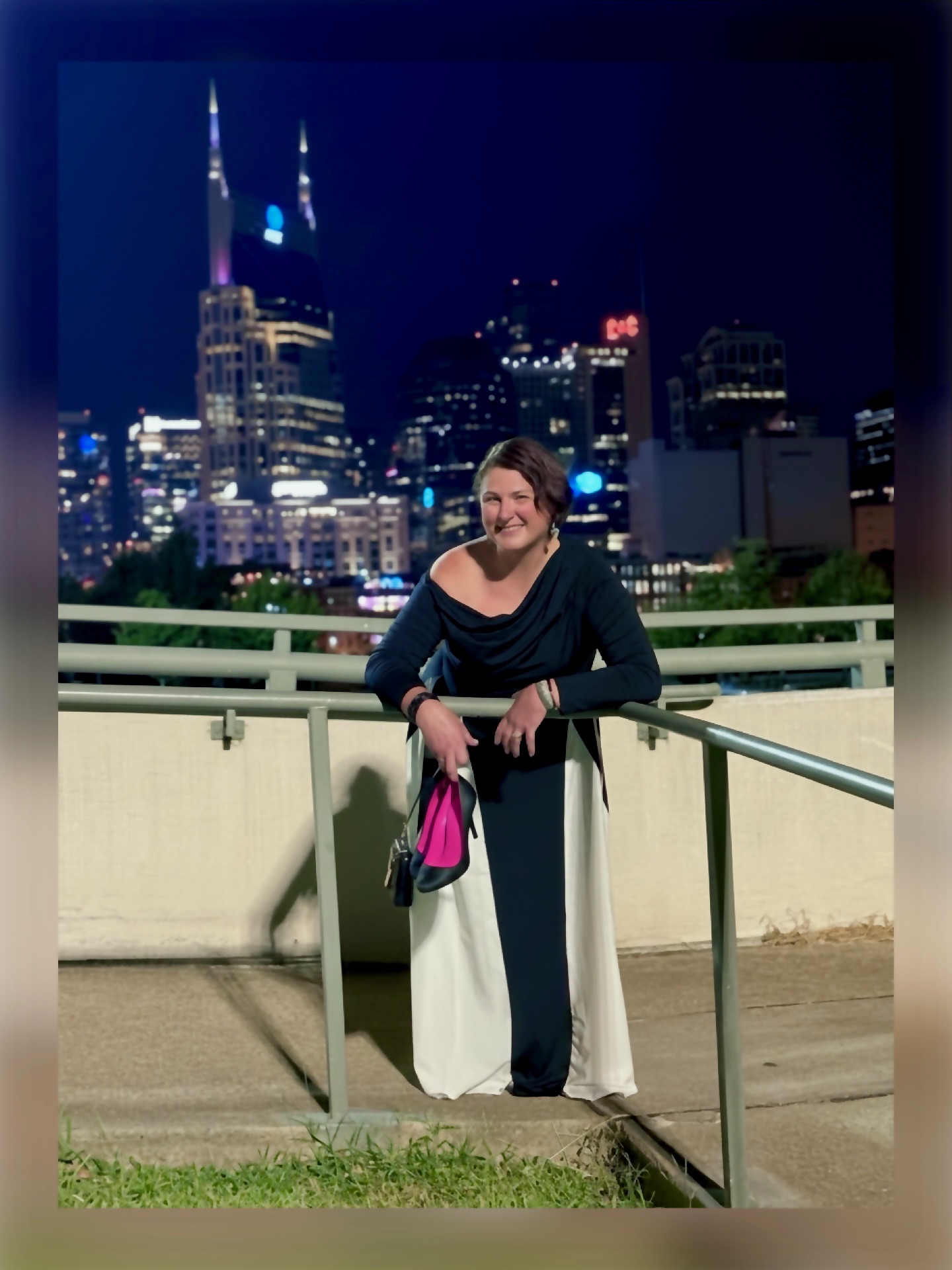 Woman standing outdoors at night with city skyline and tall buildings illuminated in the background.