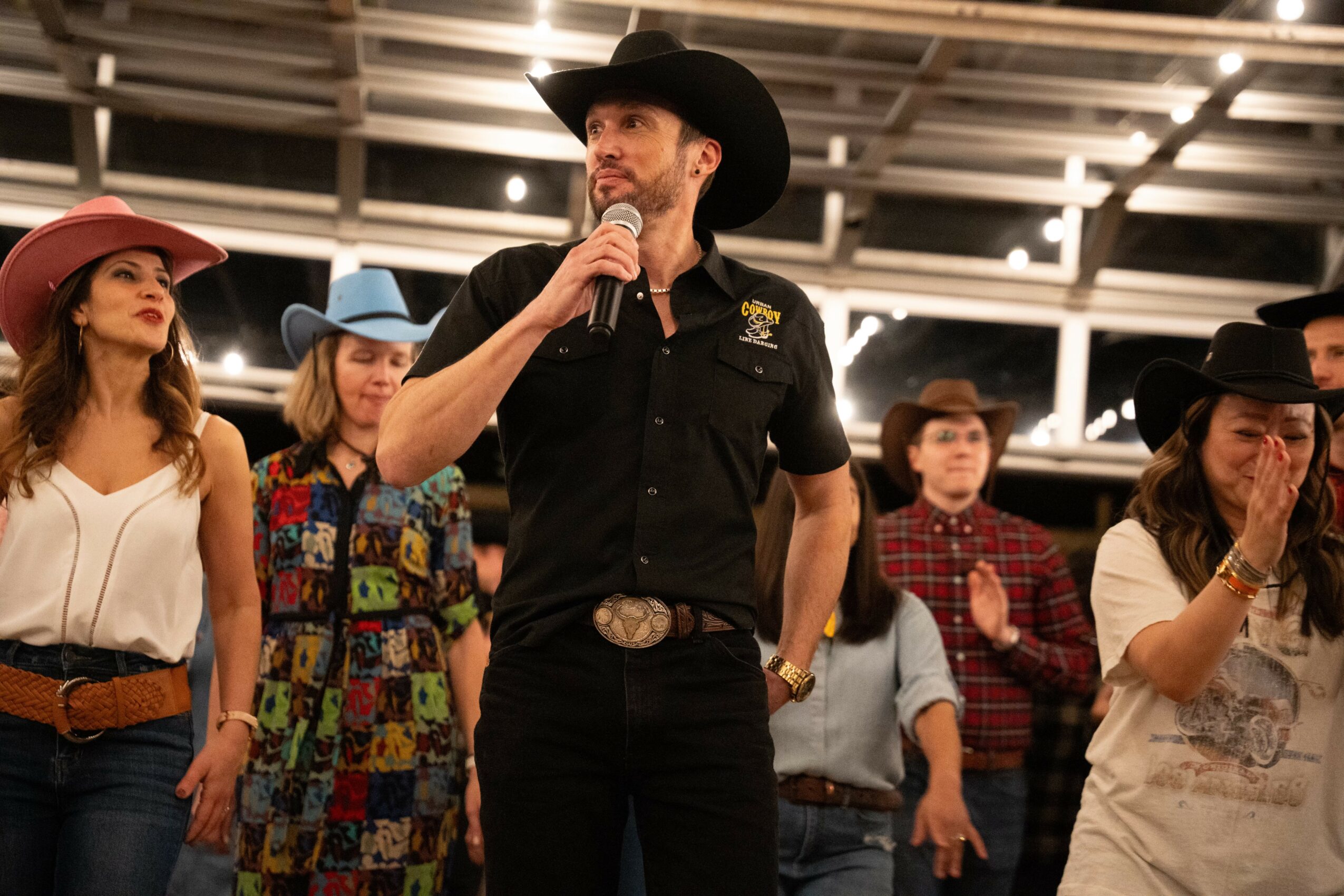 Man in black cowboy hat speaking into microphone, surrounded by women and men in cowboy hats, indoor setting with ceiling lights