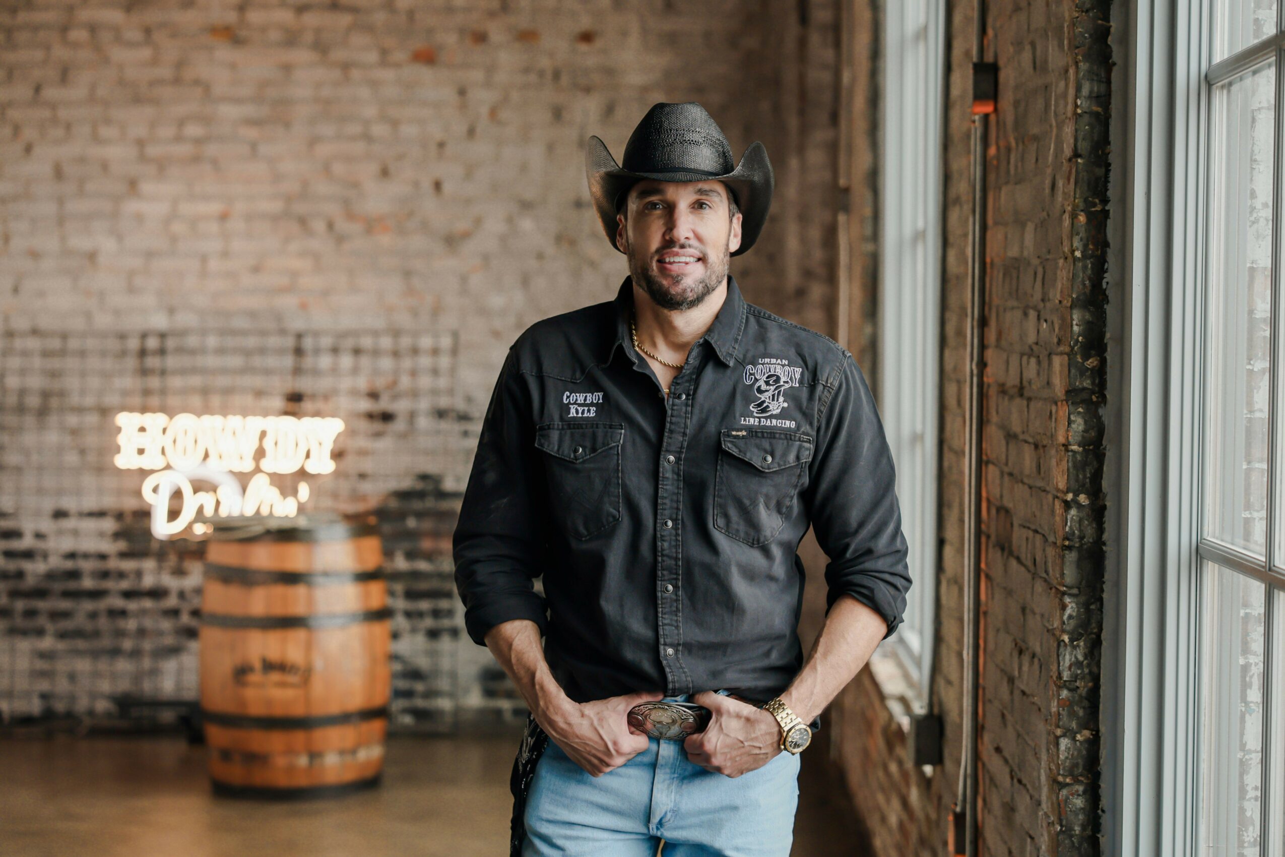 Man in black cowboy hat and black shirt standing indoors near large windows, brick wall background, smiling.