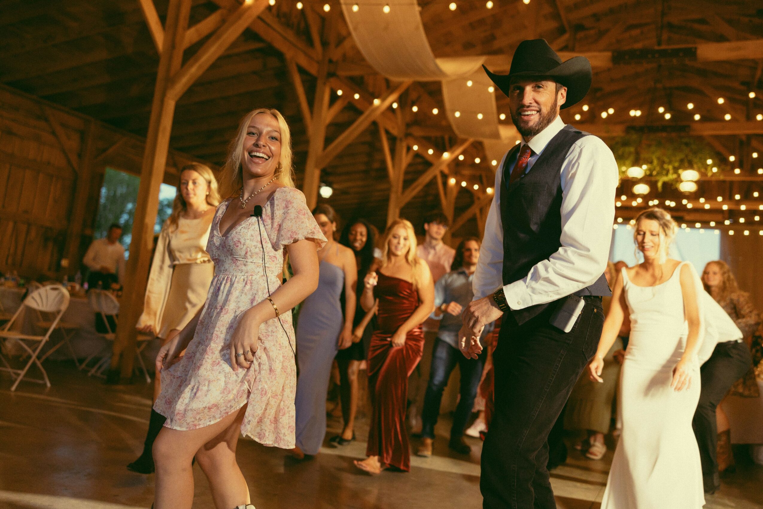 People dancing and smiling in a decorated wooden hall with string lights, including a woman in a light dress and a man in a cowboy hat.