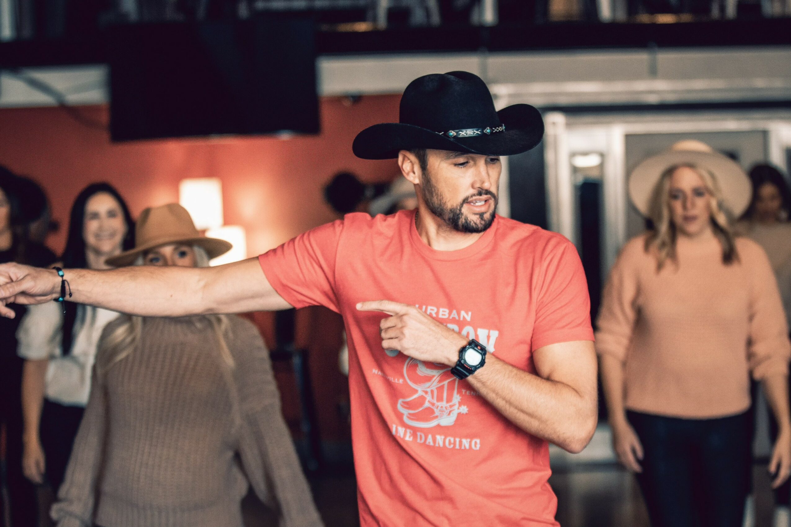 Man wearing a black cowboy hat and red T-shirt dancing in a group, with women in the background, in a lively indoor setting.