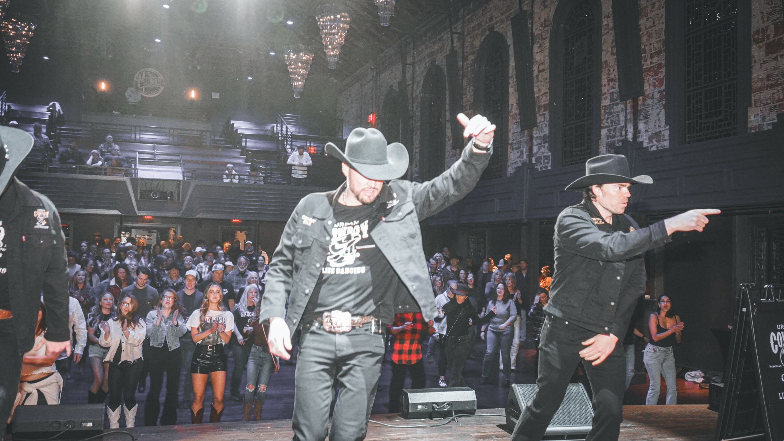 Two performers on stage with a large audience in the background, one raising an arm and the other pointing, in a concert setting.
