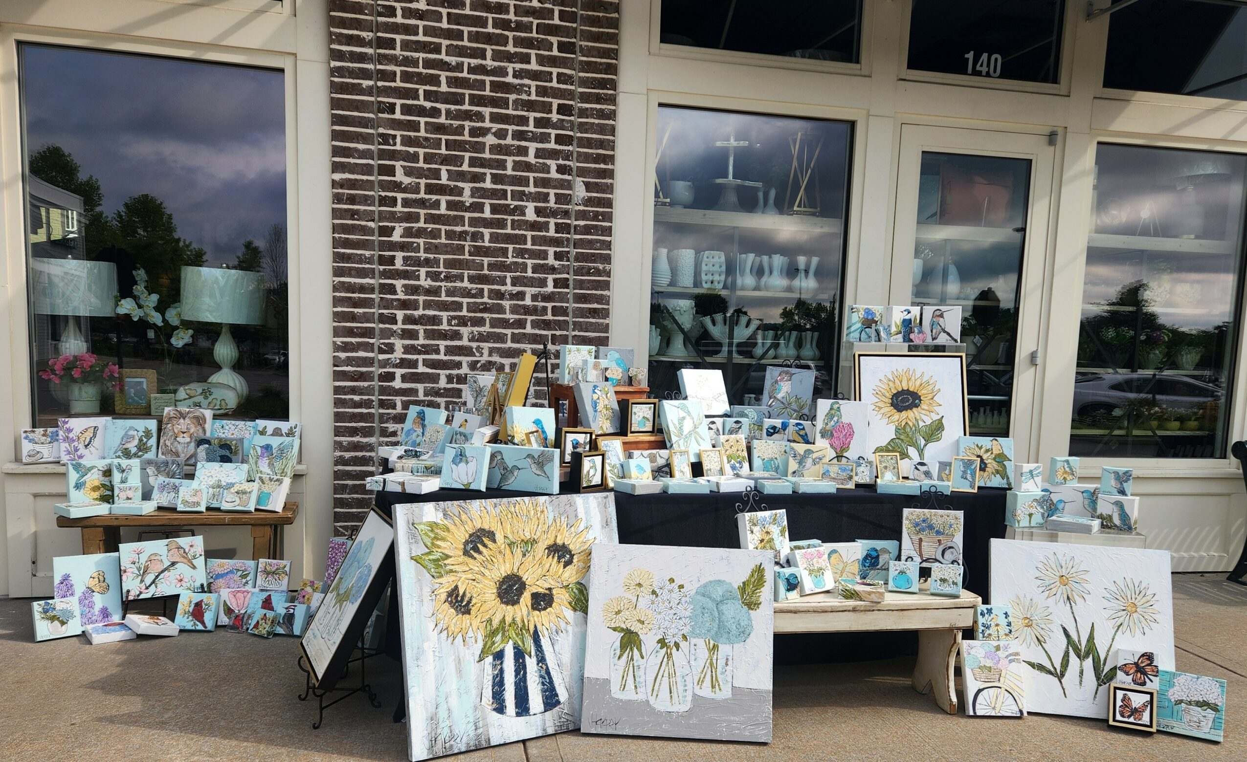 Outdoor display of paintings and artwork in front of a brick building with large windows.
