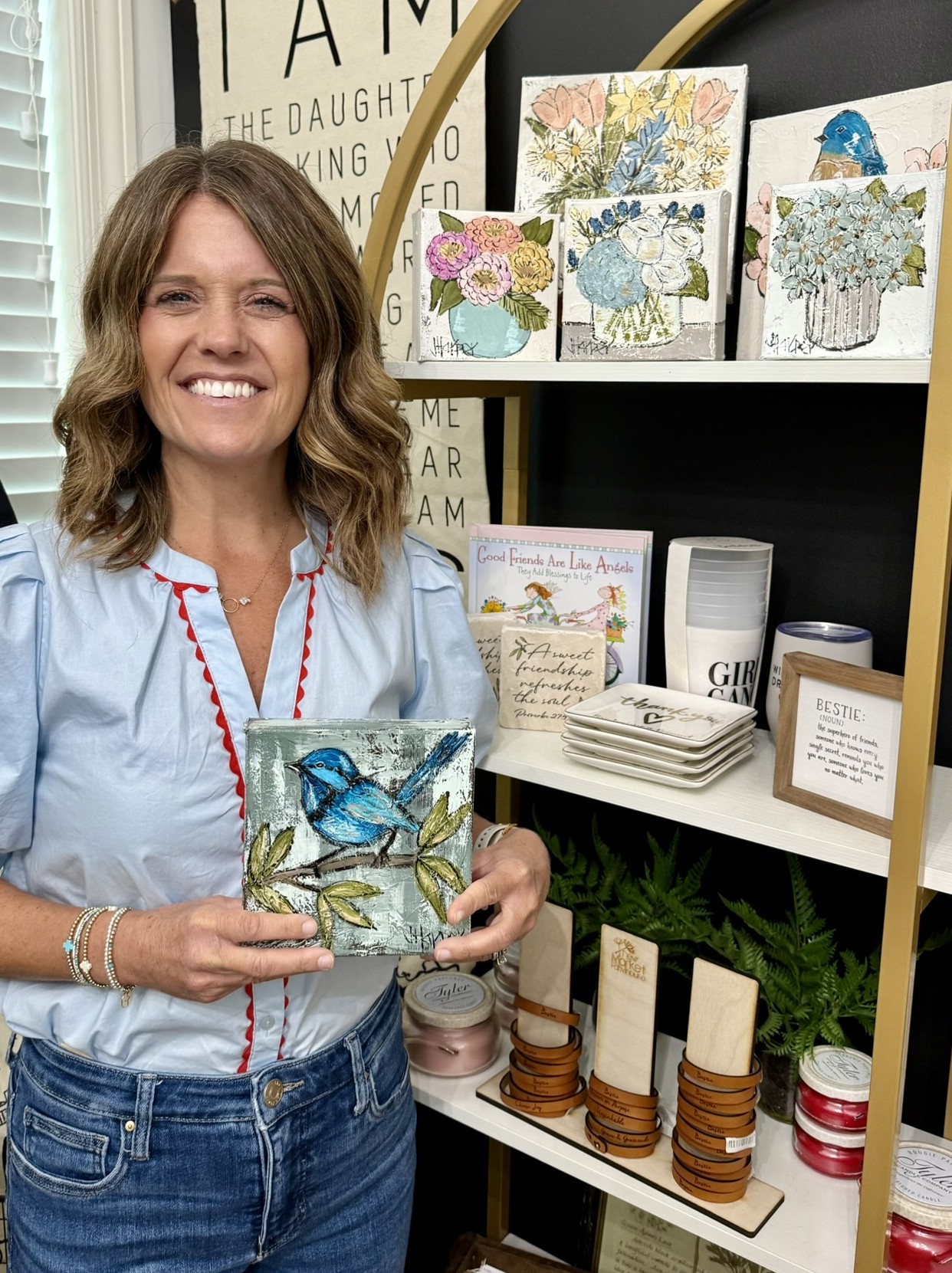 Woman smiling holding a decorative jar with a blue bird and floral design, standing in front of shelves with artwork and home decor.
