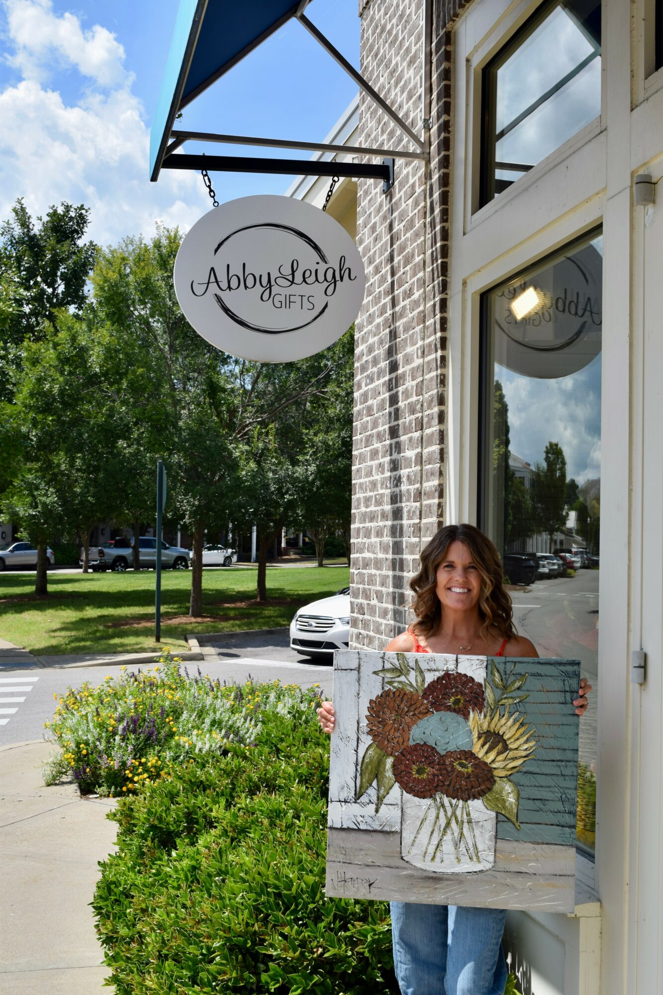 Woman holding a painted wooden sign outside a gift shop with a round sign reading 'Abby & Leigh GIFTS'.