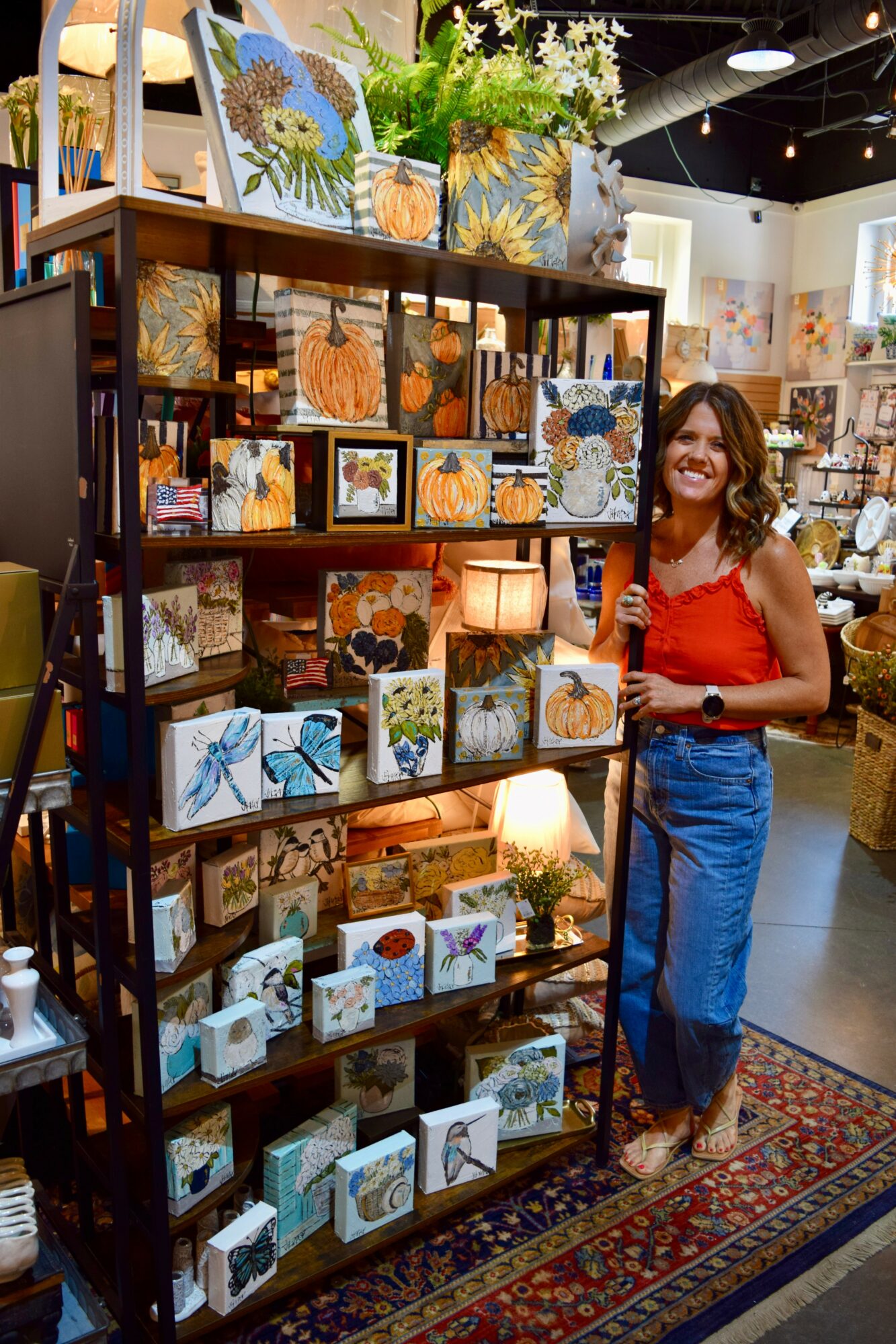 Woman in red top and blue skirt standing next to a black metal shelf filled with colorful artwork and decorative items, in an art gallery.
