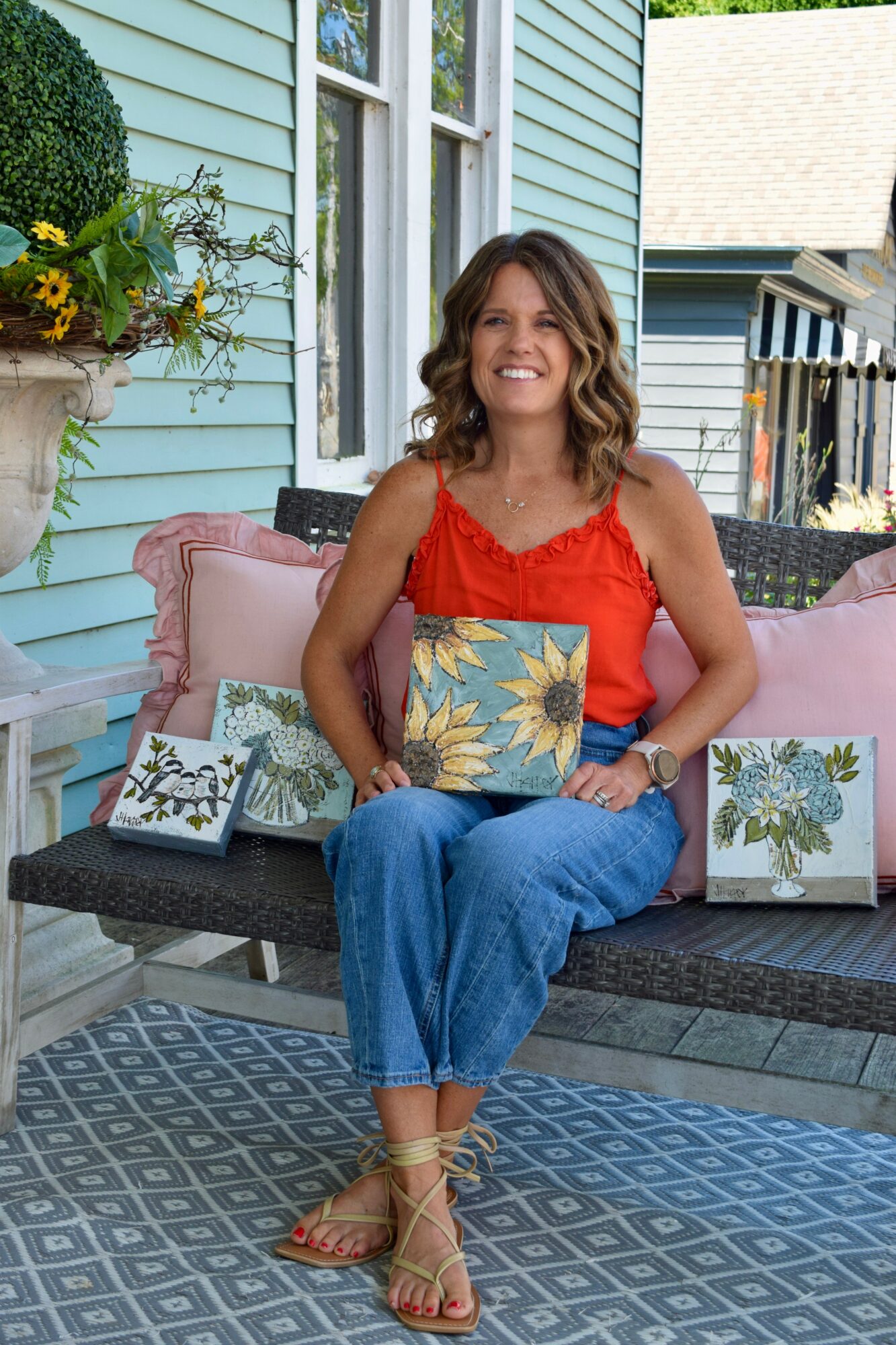 Woman sitting on a porch bench holding a floral pillow, smiling, with decorative pillows and plants around her.