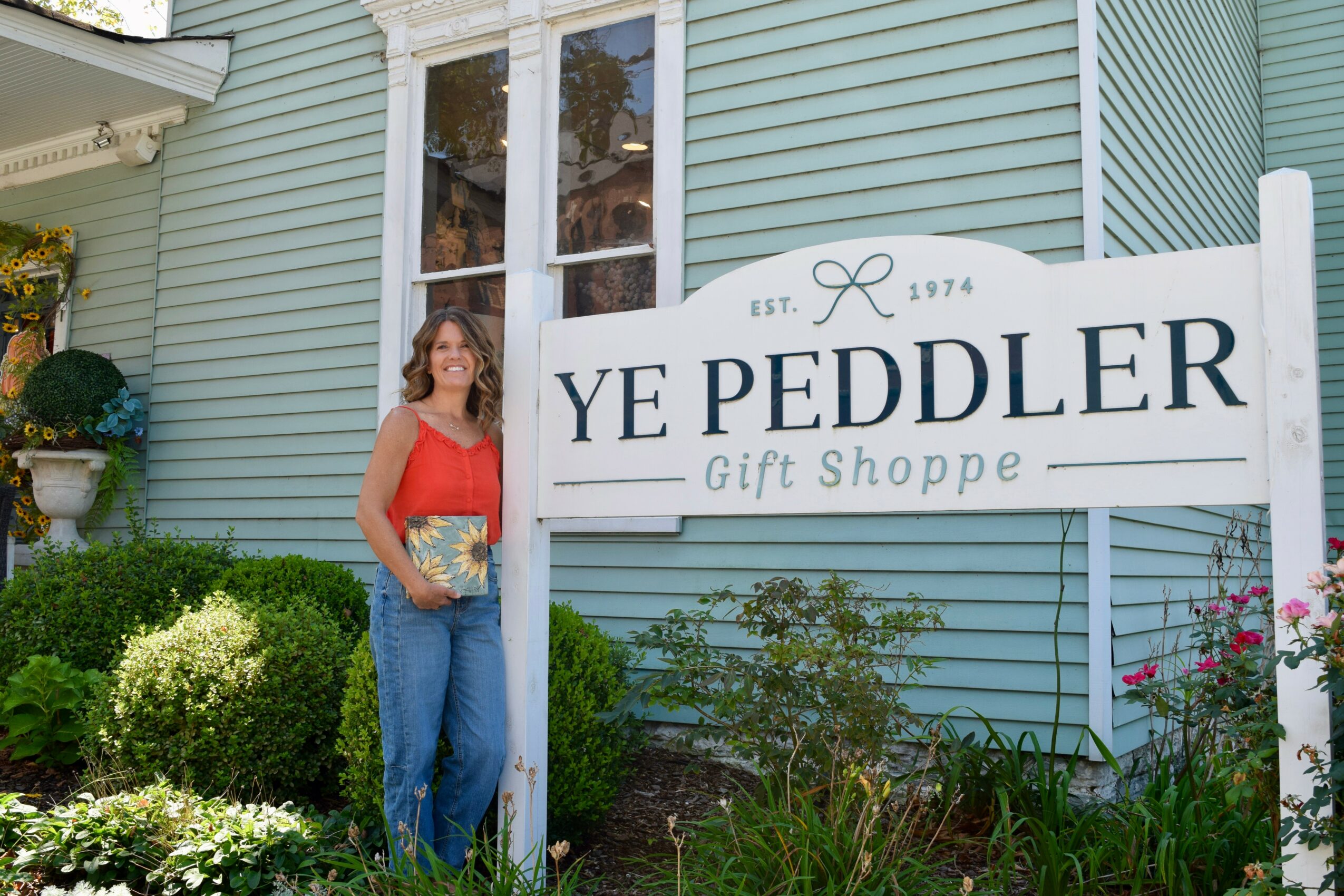 Woman standing next to a sign for Ye Peddler Gift Shoppe outside a light blue building, holding a gift box.