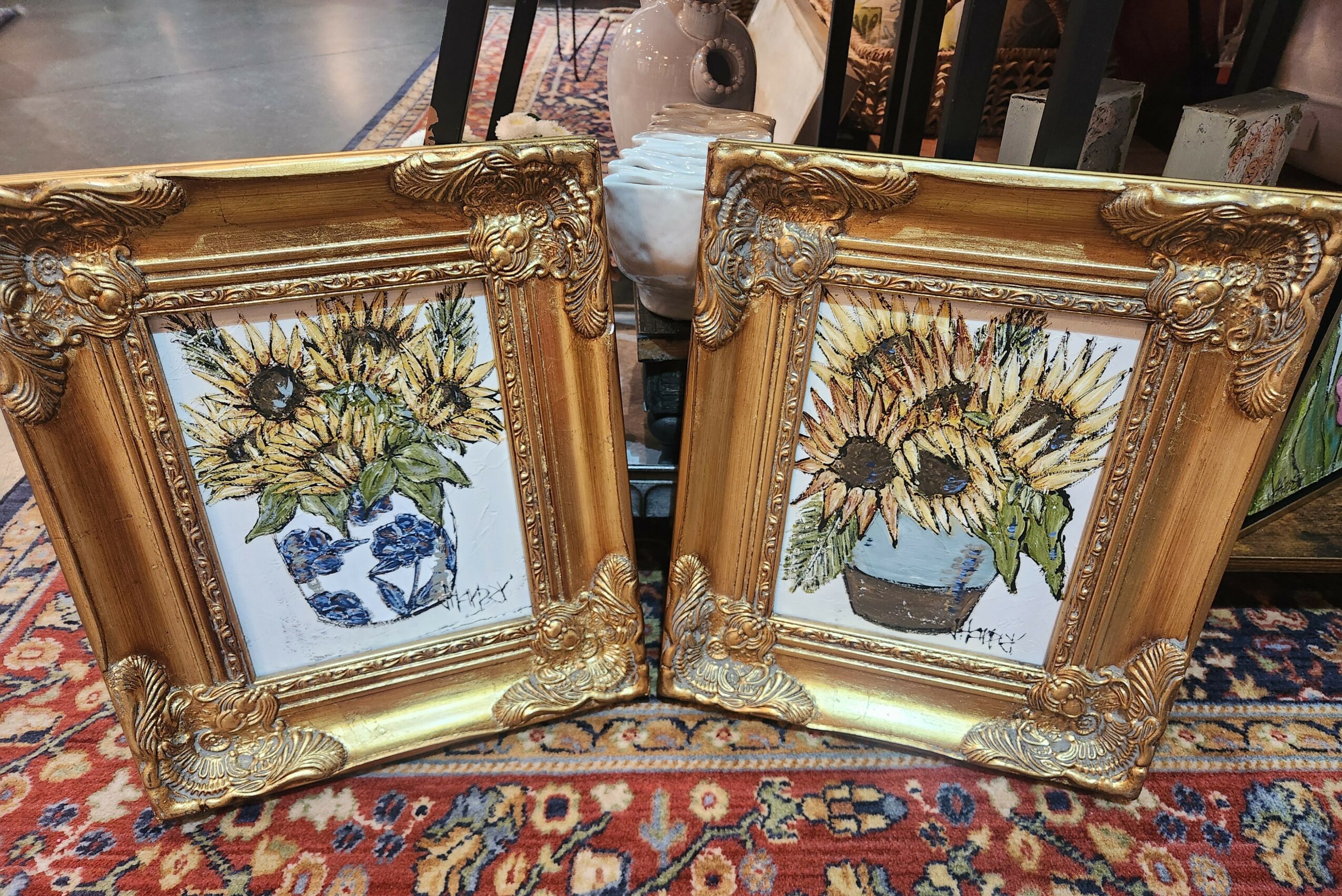 Two framed sunflower paintings in ornate gold frames, placed on a patterned rug. One painting shows a blue and white vase, the other a brown pot.