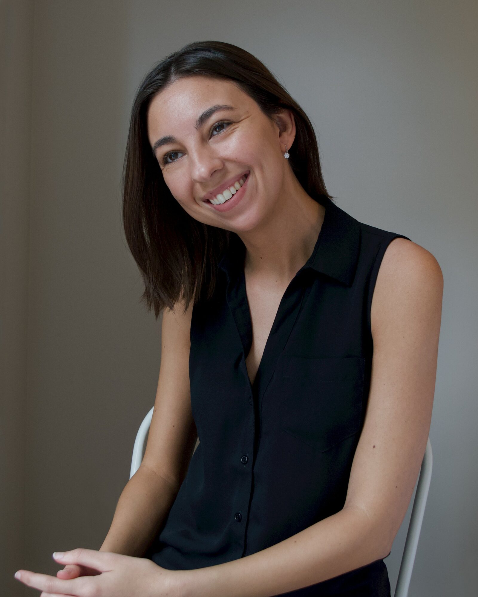 Smiling woman with shoulder-length dark hair wearing a sleeveless black top, sitting indoors.