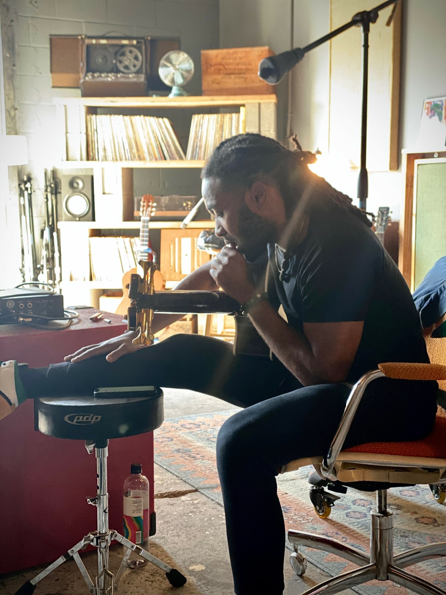 Person with long hair sitting in a room, playing a harmonica, surrounded by books, music equipment, and a microphone.