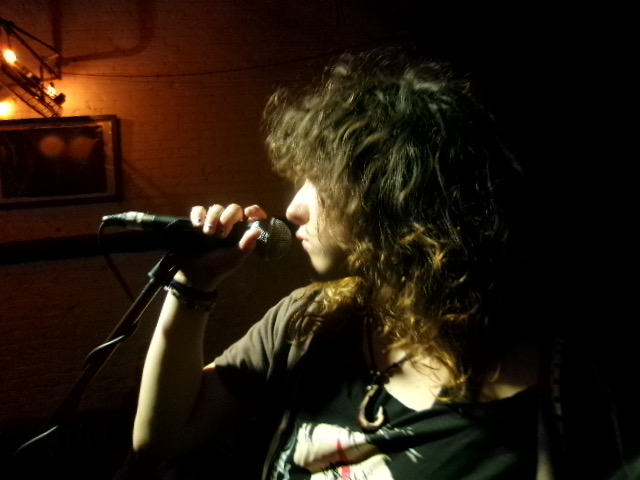 Person with curly hair holding a microphone, facing left, in a dimly lit setting.