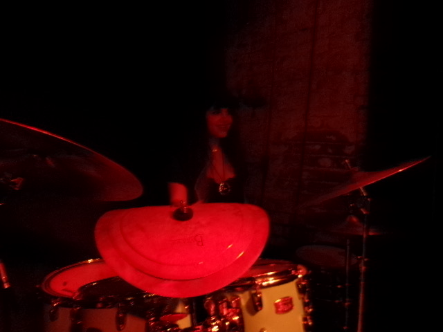 Person playing drums in a dark setting with red lighting, partially obscured by drum equipment.