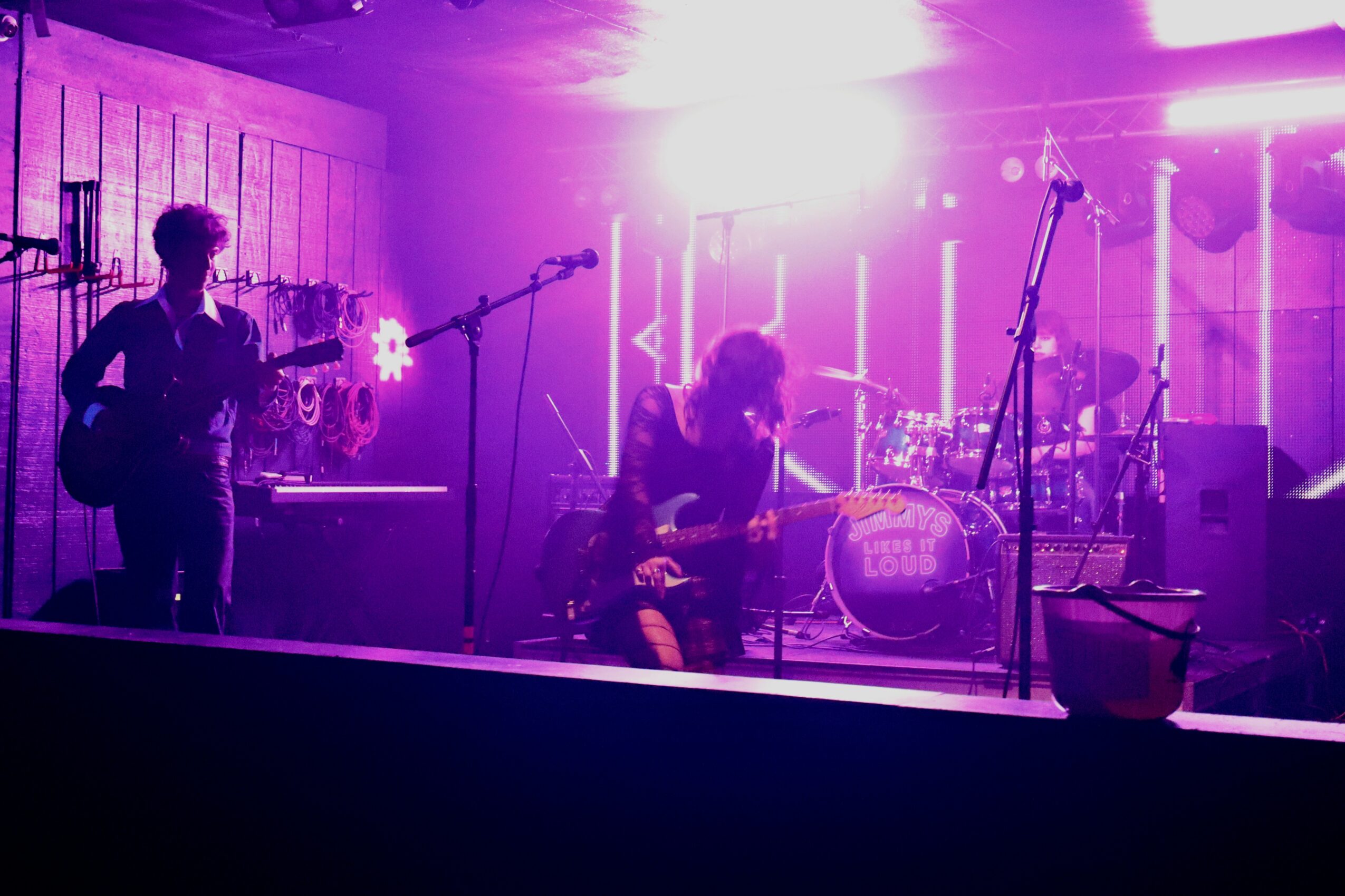 Band performing on stage with purple lighting, two guitarists and a drummer visible, microphones and equipment present.