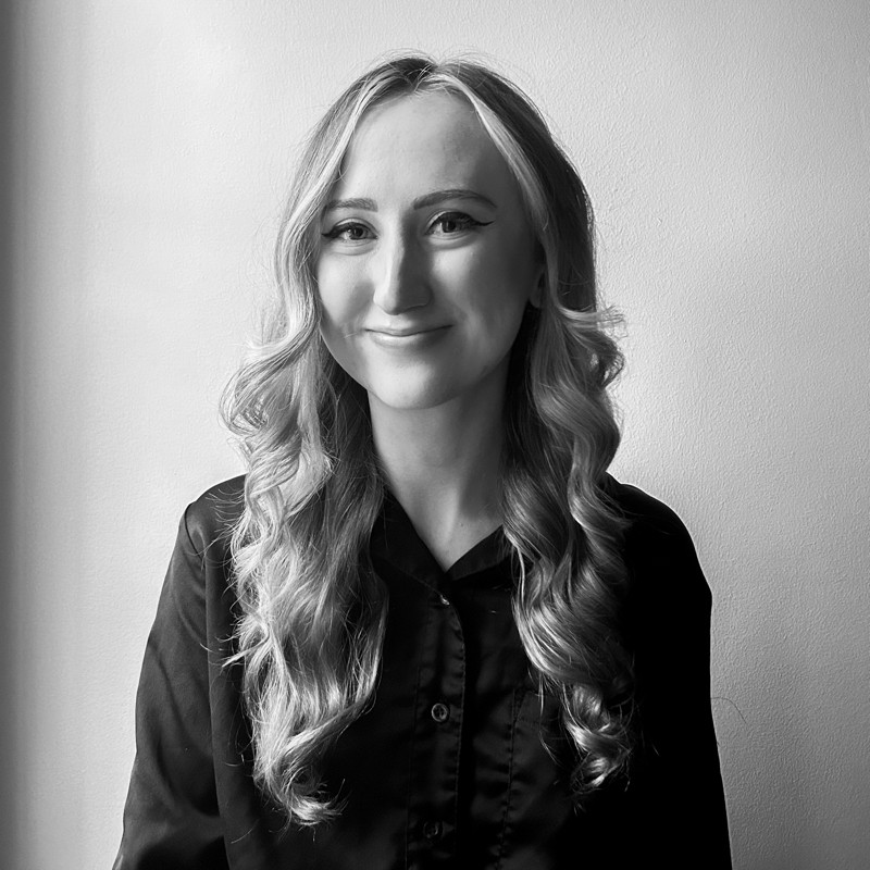Young woman with long wavy hair smiling, wearing a dark shirt, standing against a plain wall.
