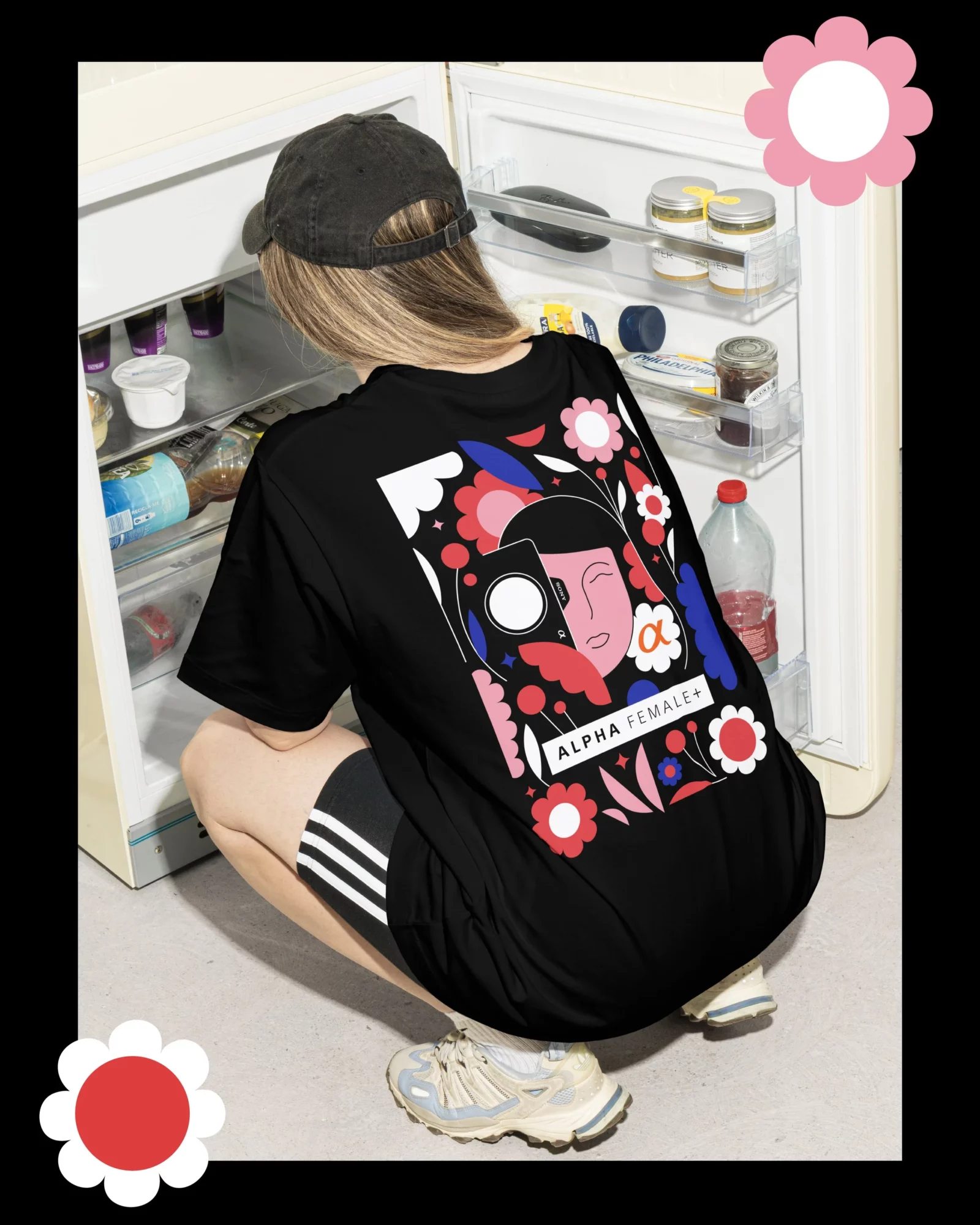 Person crouching in front of an open refrigerator, wearing a black cap and graphic t-shirt with colorful design, shorts, and sneakers.