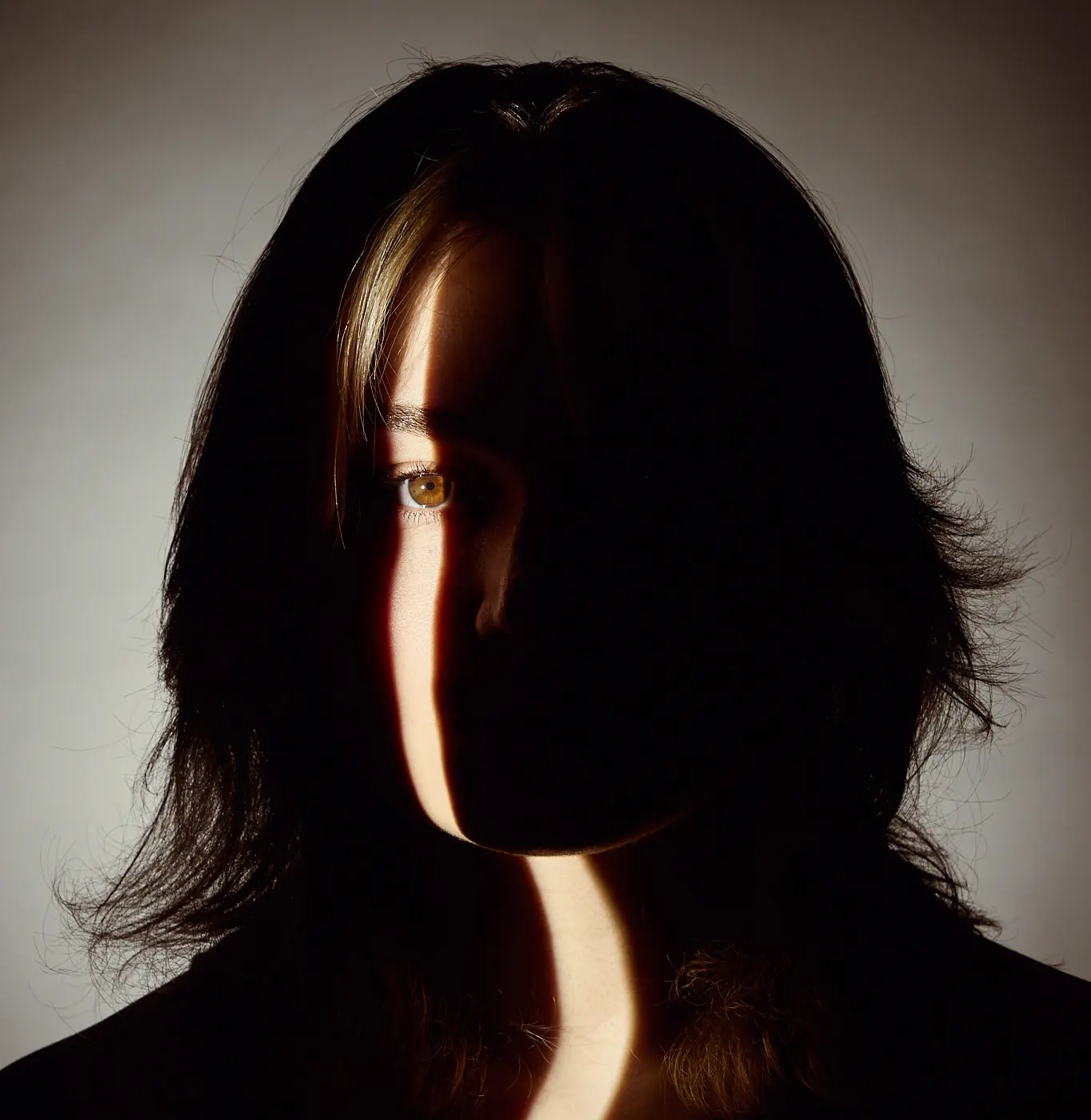 Person with dark hair partially illuminated by a vertical light streak on face, dark background.