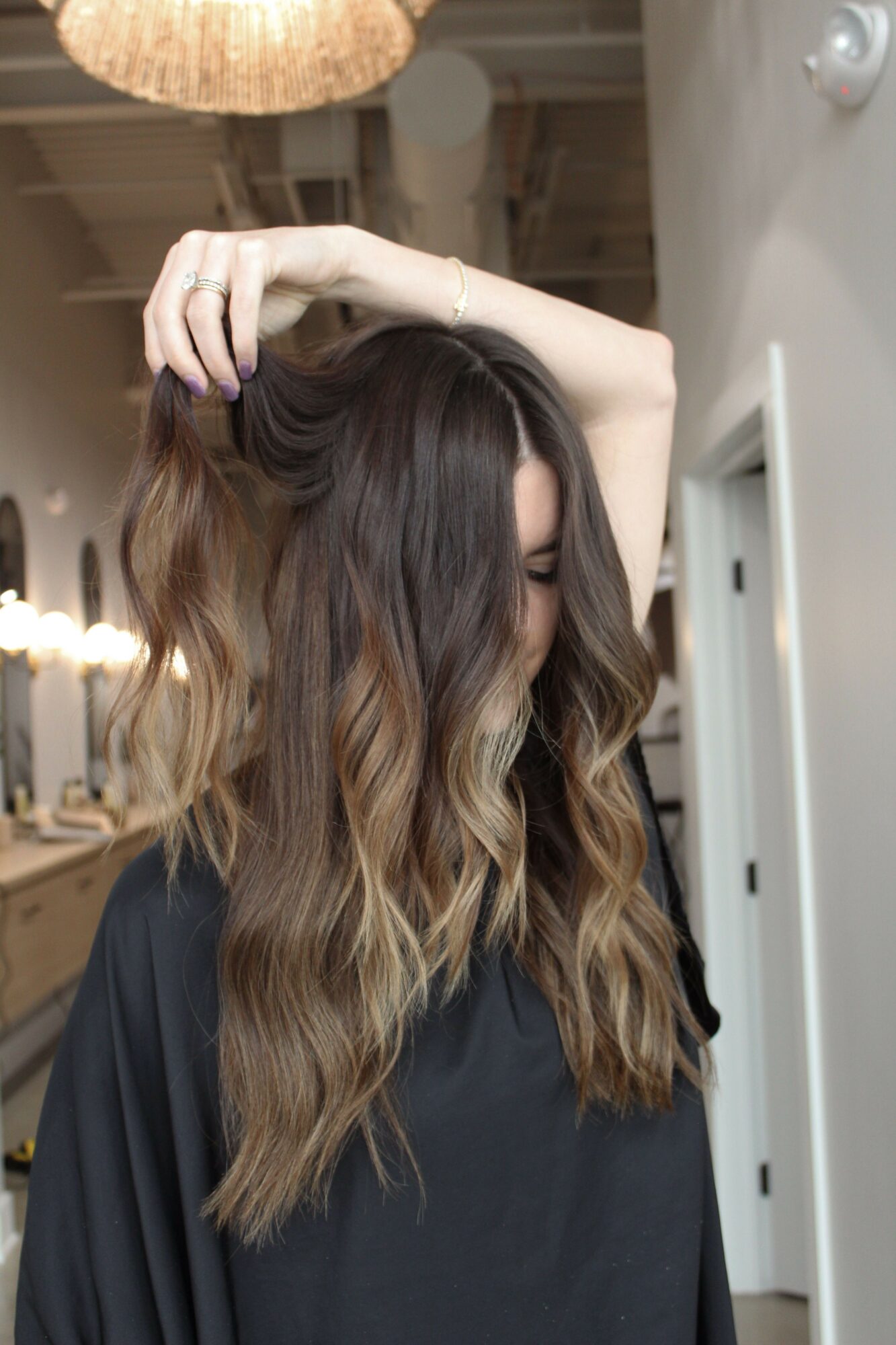 Person with long, wavy hair covering face, hand touching hair, in indoor setting with warm lighting.