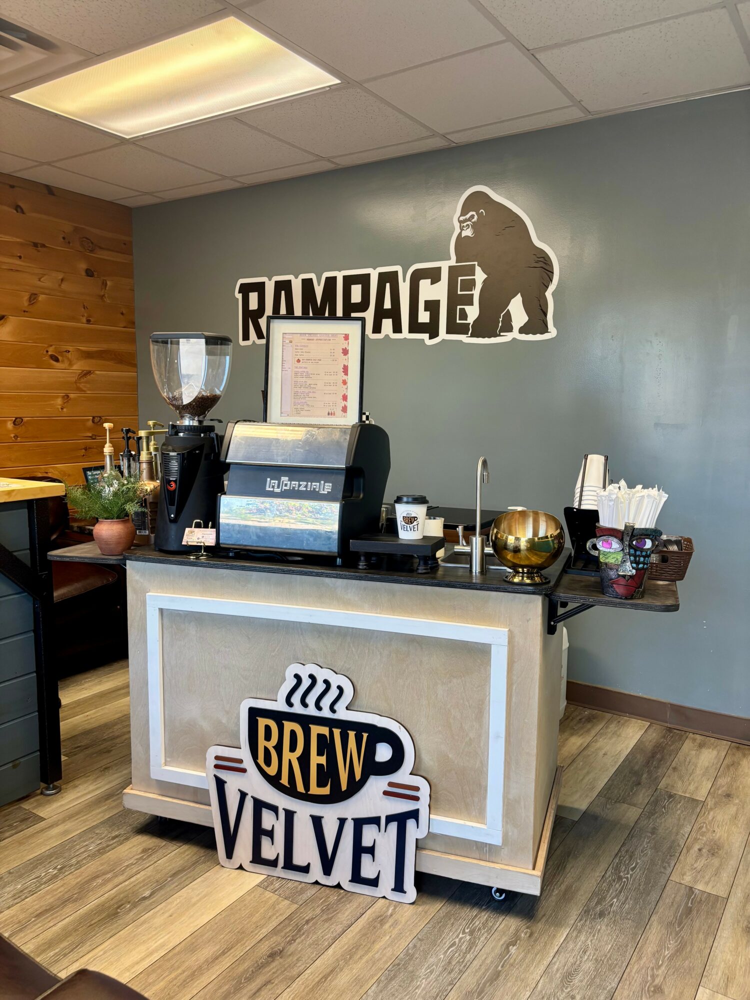 Coffee shop counter with a sign reading 'Brew Velvet' and a wall sign with a gorilla silhouette and 'RAMPAGE' text, wooden and gray decor.
