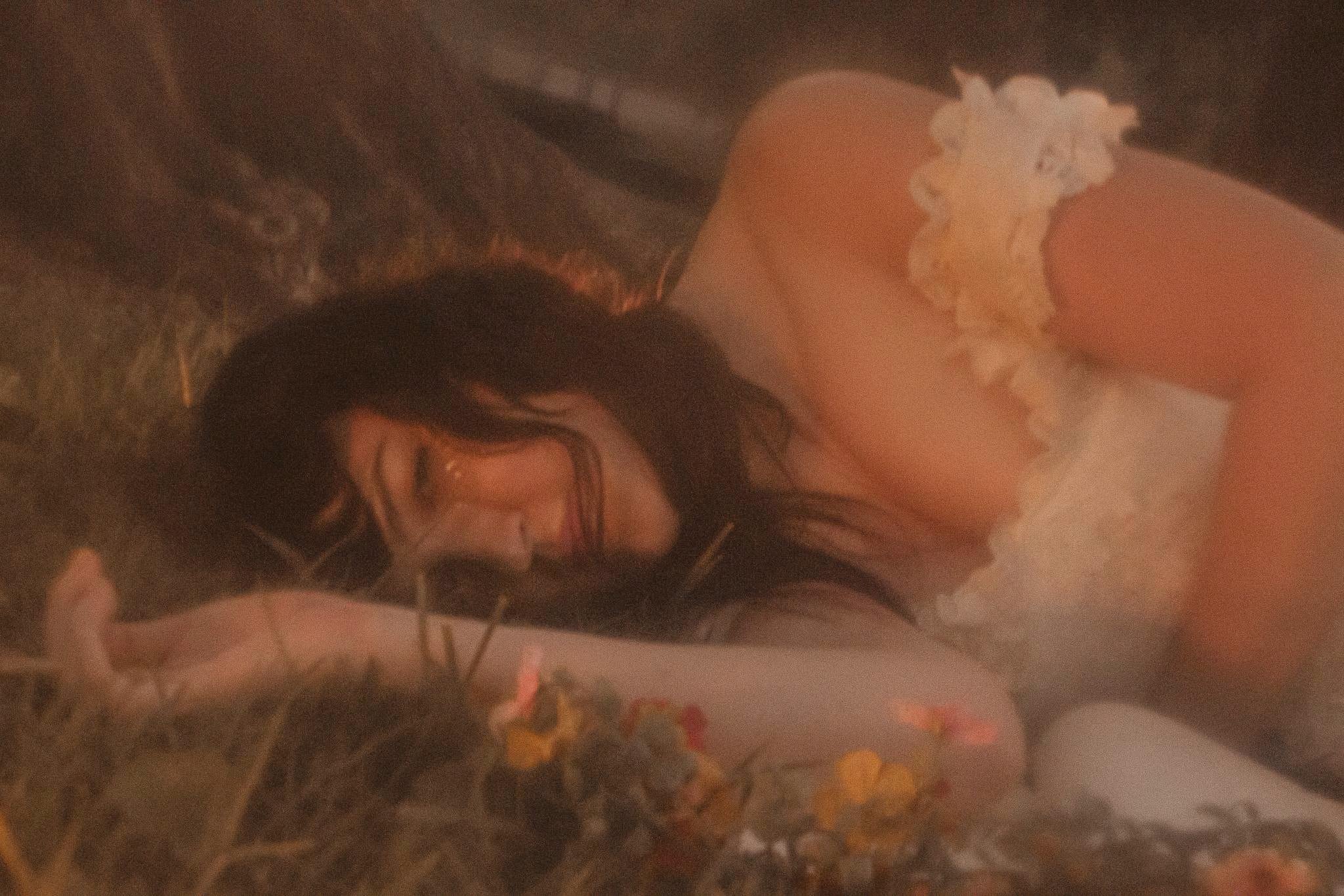 Woman lying on grass with flowers, resting her head on her arm, smiling, wearing a light-colored dress with lace details.